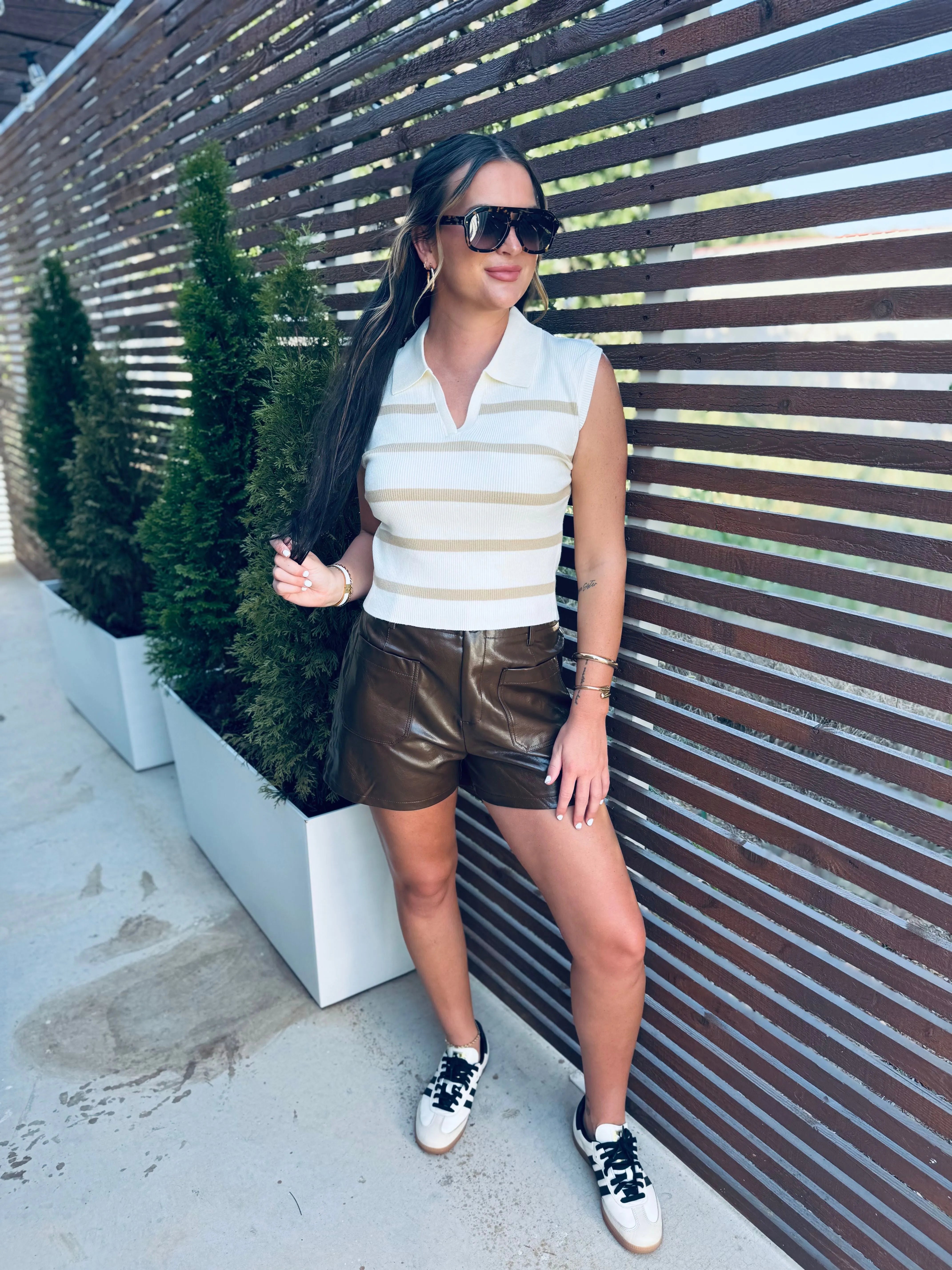 Woman wearing a cream and gold striped polo vest, brown leather shorts, and black and white sneakers, standing outdoors.