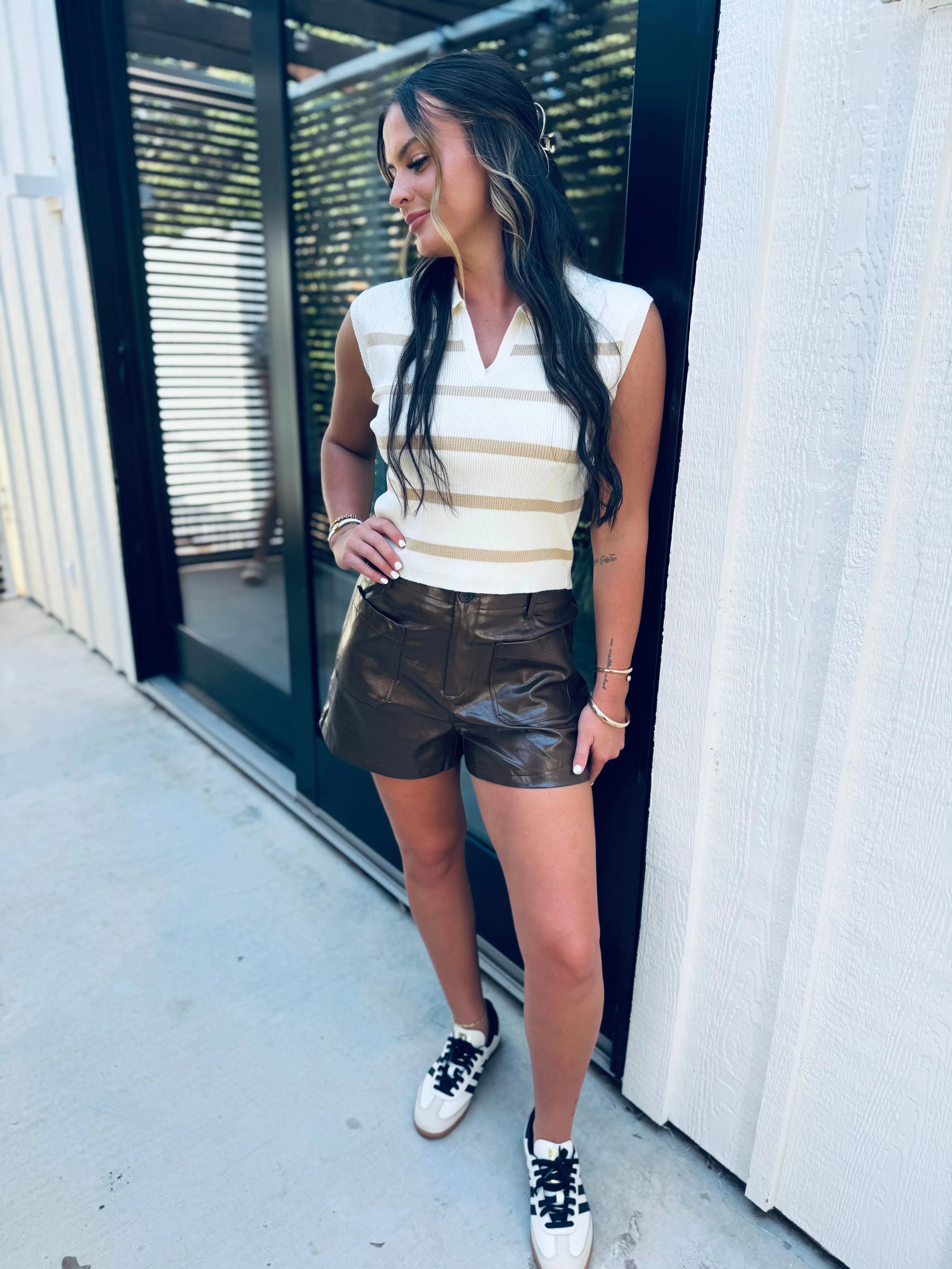Woman wearing a sleeveless cream and gold striped polo vest with a v-neck, paired with dark brown faux leather shorts and