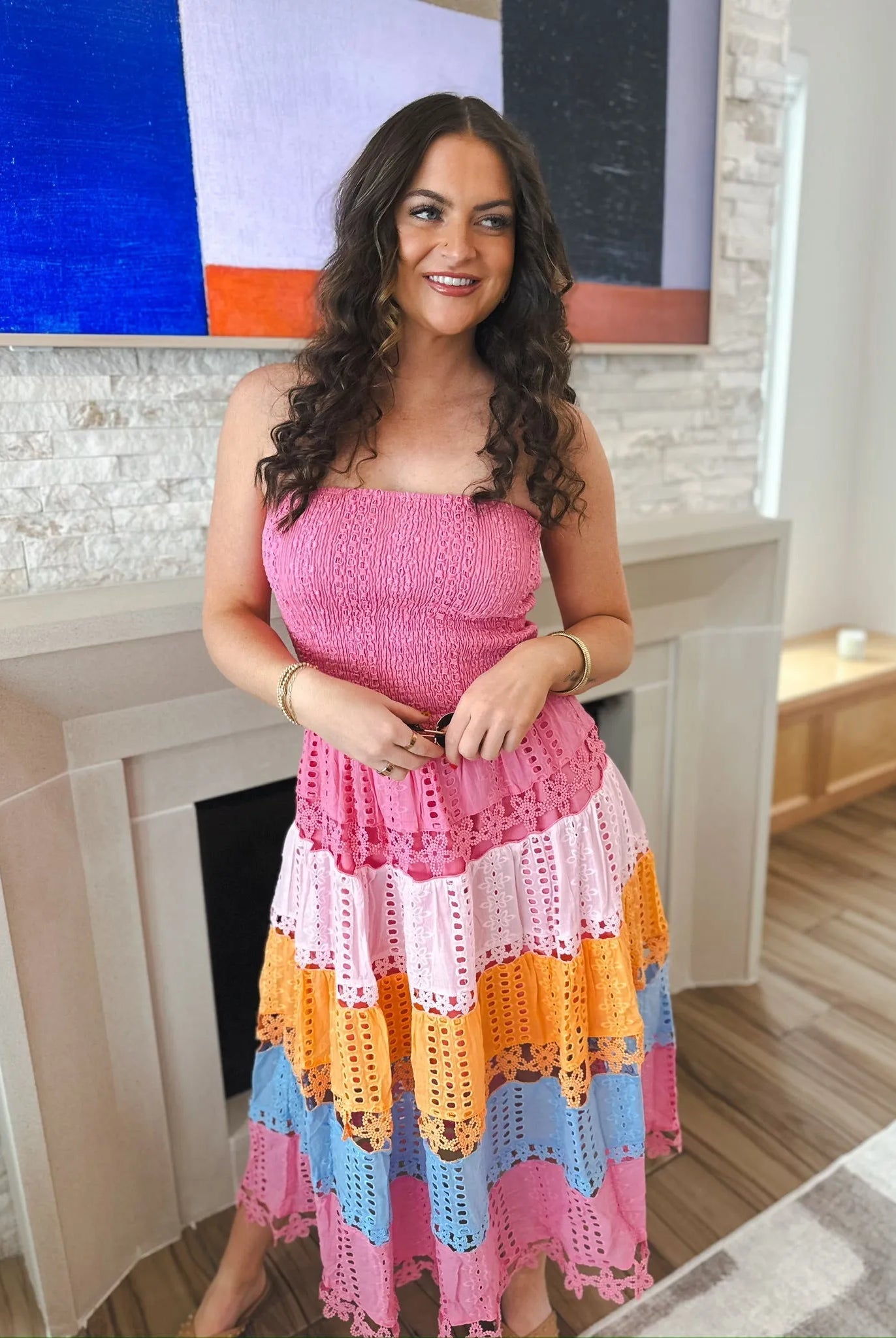 Woman wearing a pink strapless midi dress with eyelet lace tiers in pink, light pink, orange, and blue. She has curly brown