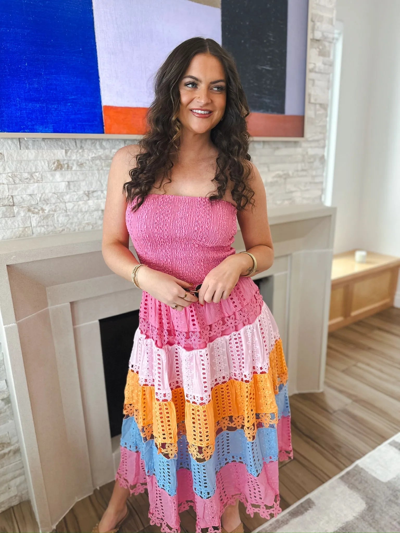 Woman wearing a pink strapless midi dress with eyelet lace tiers in pink, light pink, orange, and blue. She has curly brown