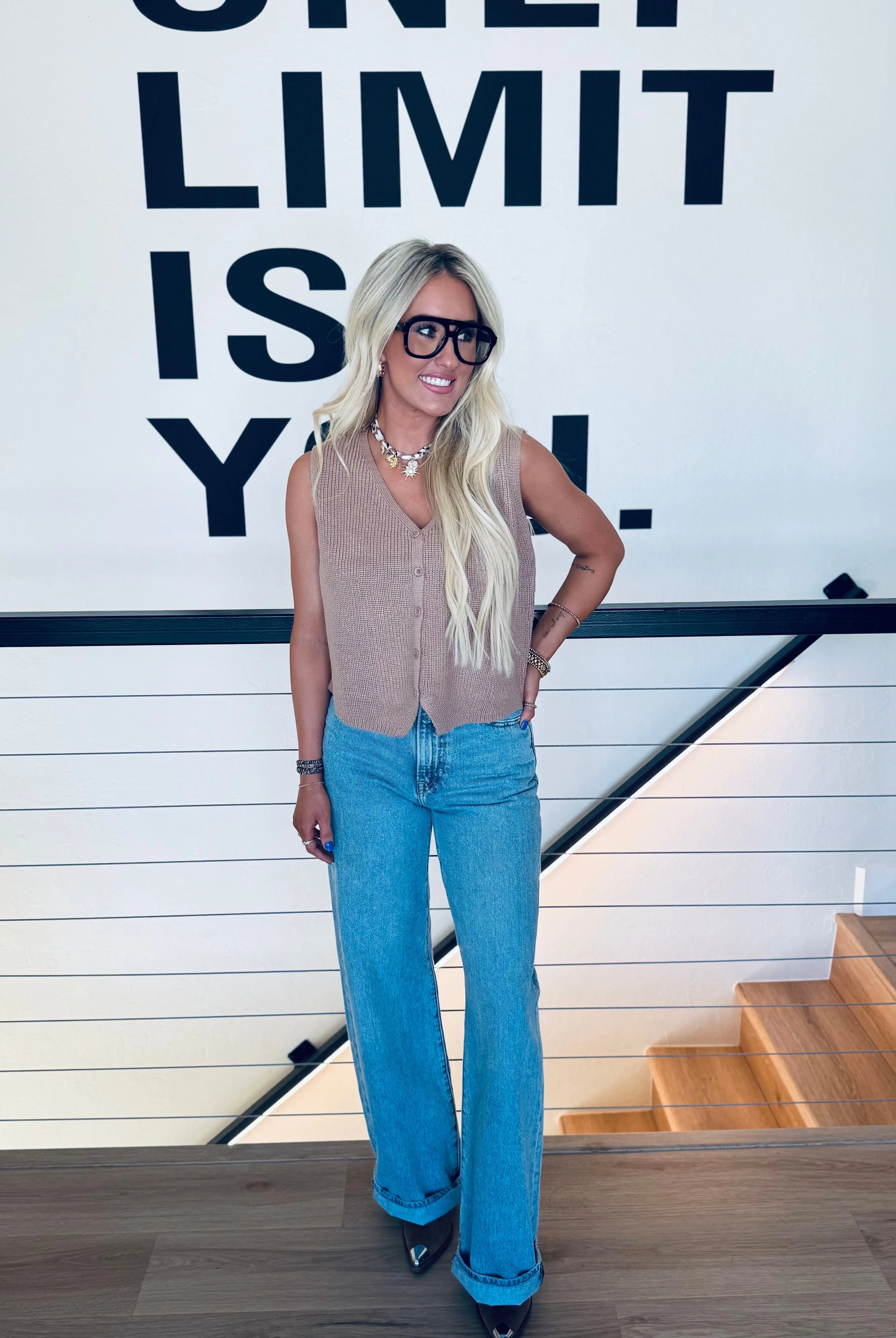 Woman wearing a taupe button-down sweater vest and light wash wide-leg jeans, accessorized with large black glasses and