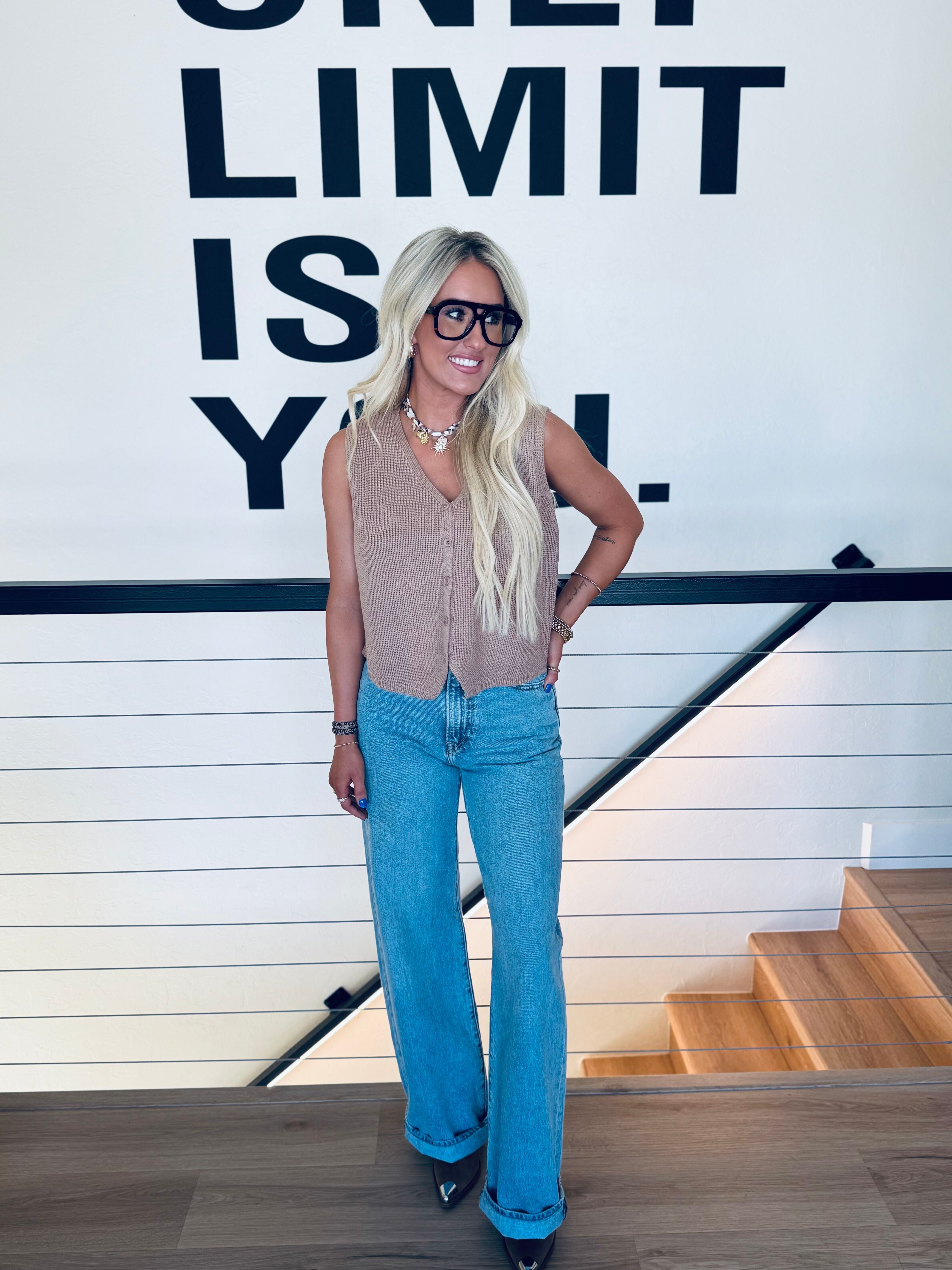 Woman wearing a taupe button-down sweater vest and light wash wide-leg jeans, accessorized with large black glasses and