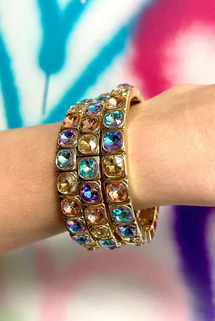 Three stacked gold bracelets adorned with colorful cushion-cut crystals in blue, purple, pink, and amber tones.
