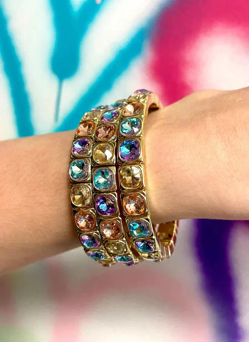 Three stacked gold bracelets adorned with colorful cushion-cut crystals in blue, purple, pink, and amber tones.