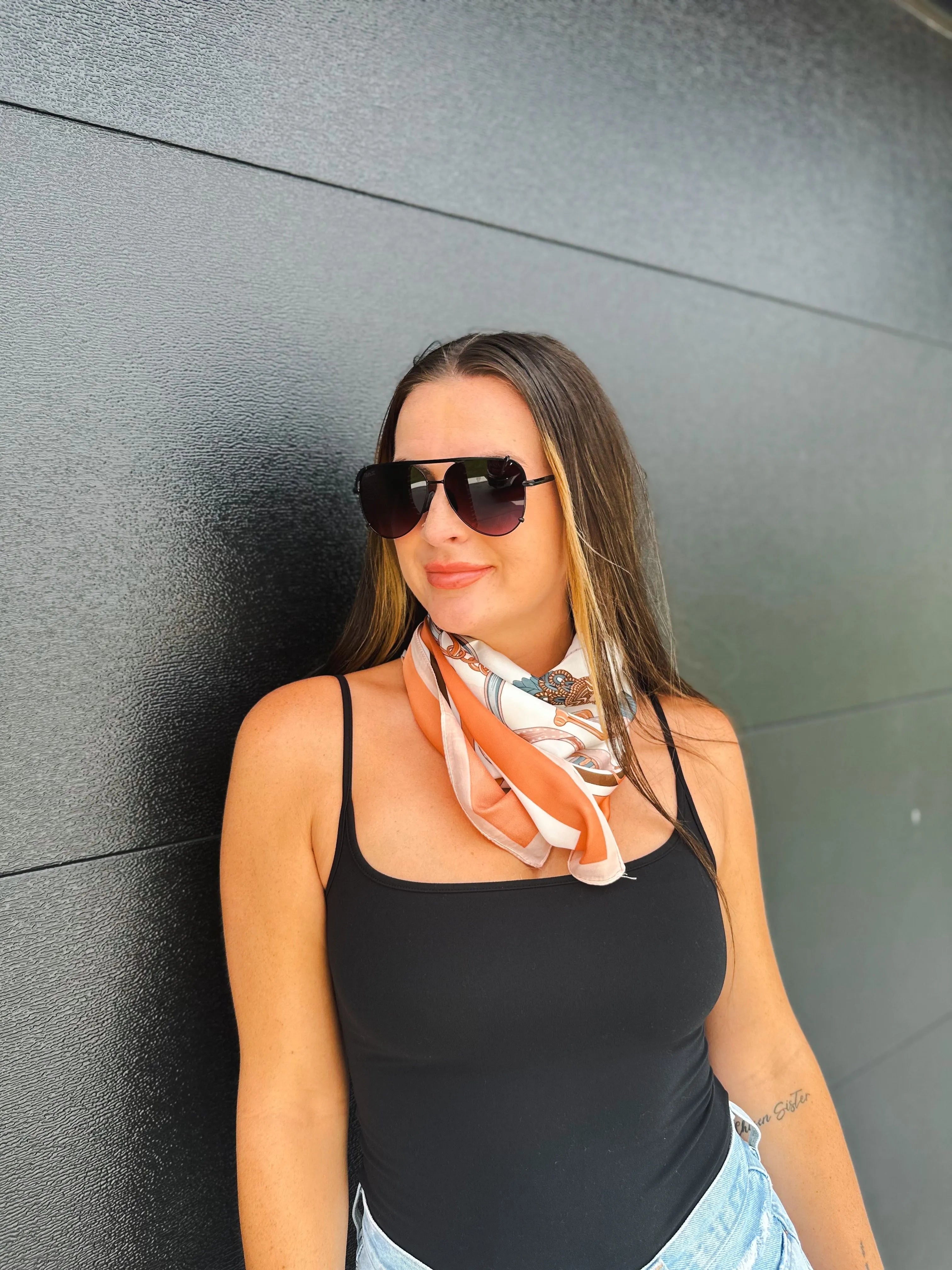 Woman wearing a black tank top and a teal orange rope print neck scarf with white accents. She is wearing aviator sunglasses.