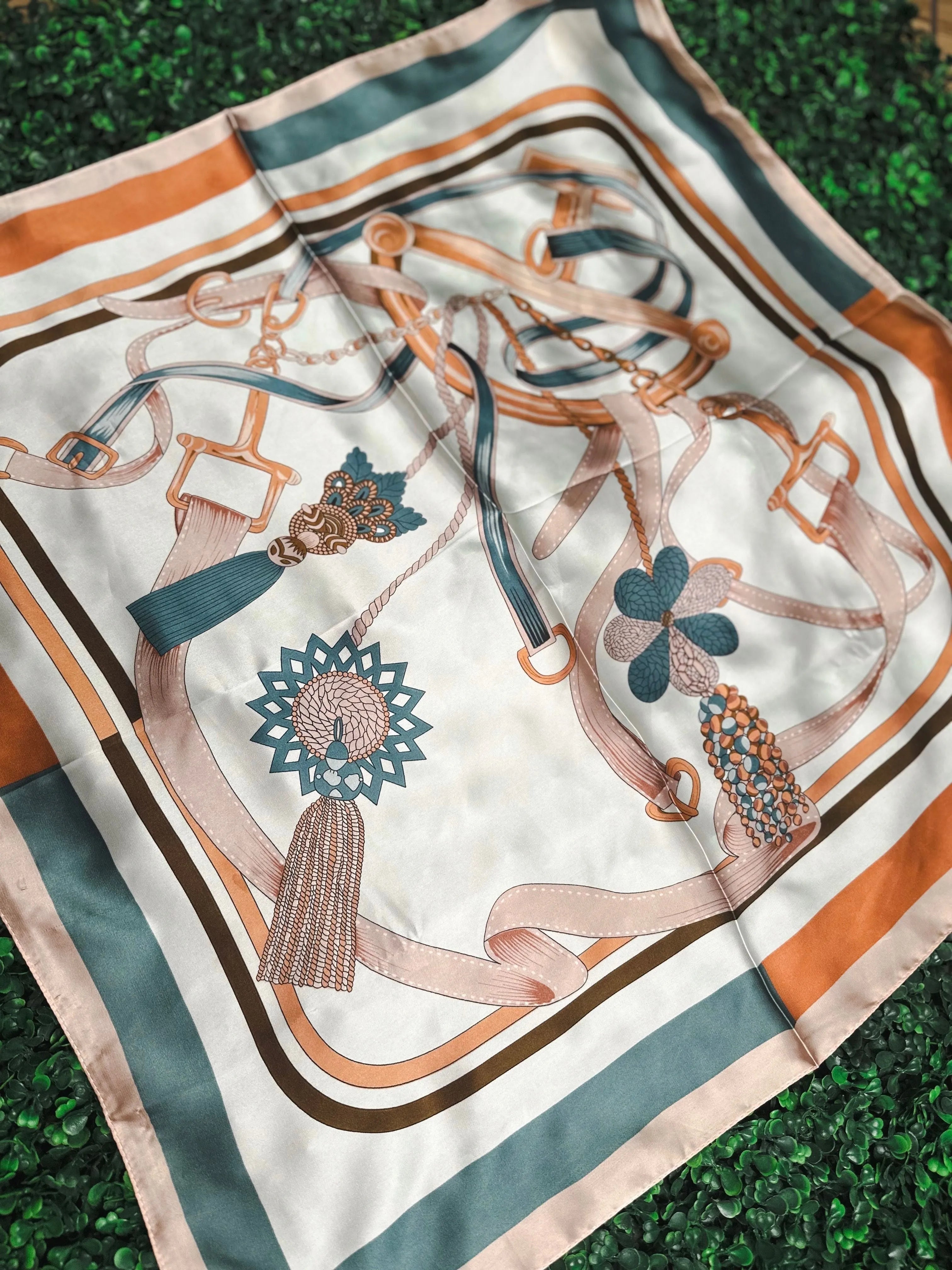 White silk scarf with a print of teal, orange, and peach belts, chains, and tassels. Bordered with stripes of teal, brown