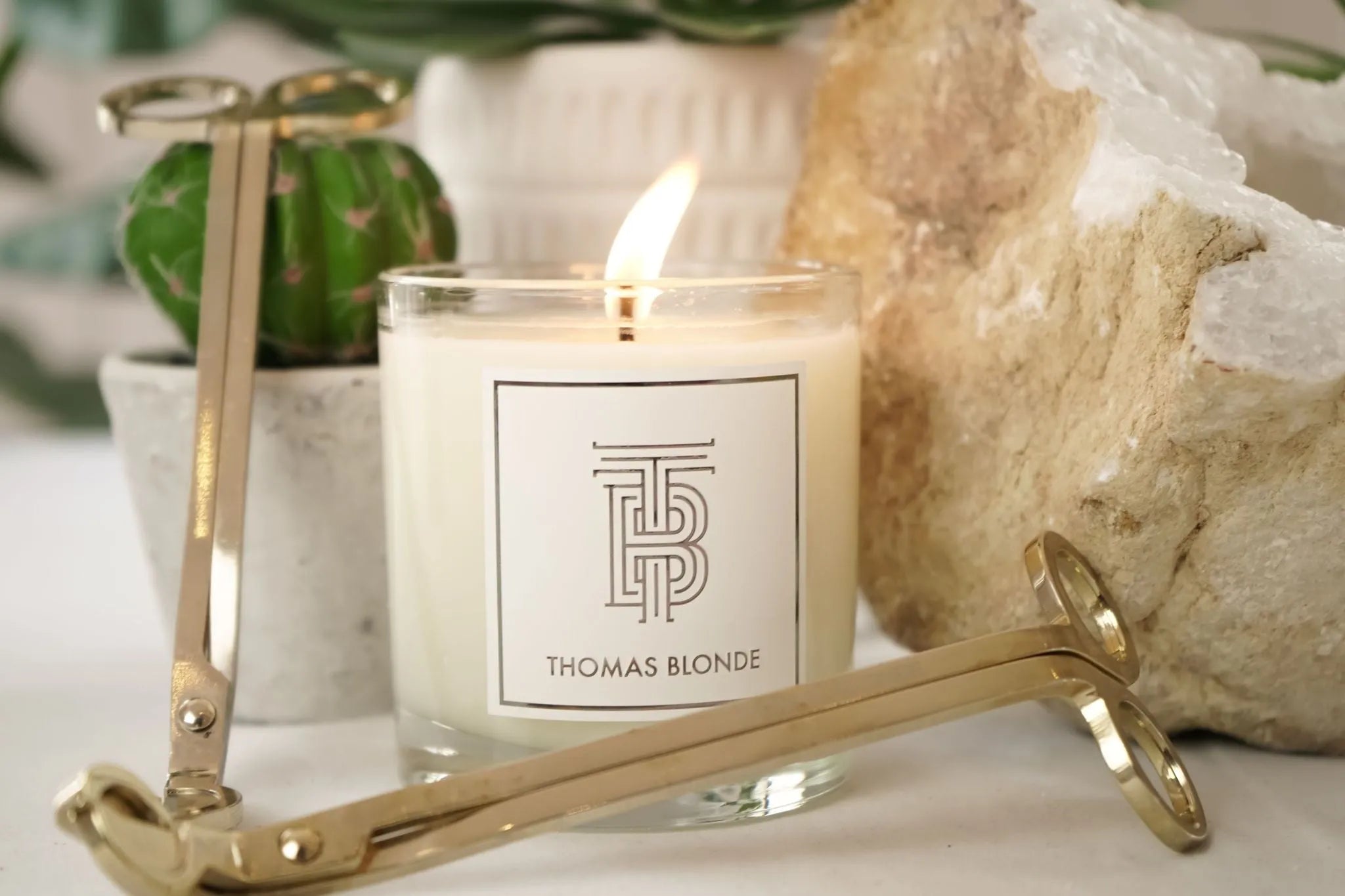 Thomas Blonde signature soy wax candle in a clear glass jar with a lit wick. Gold wick trimmers surround the candle on a