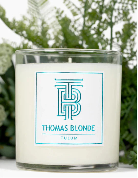 Thomas Blonde Tulum candle in a glass jar with a white label featuring teal "TB" logo and text. Green foliage in background.