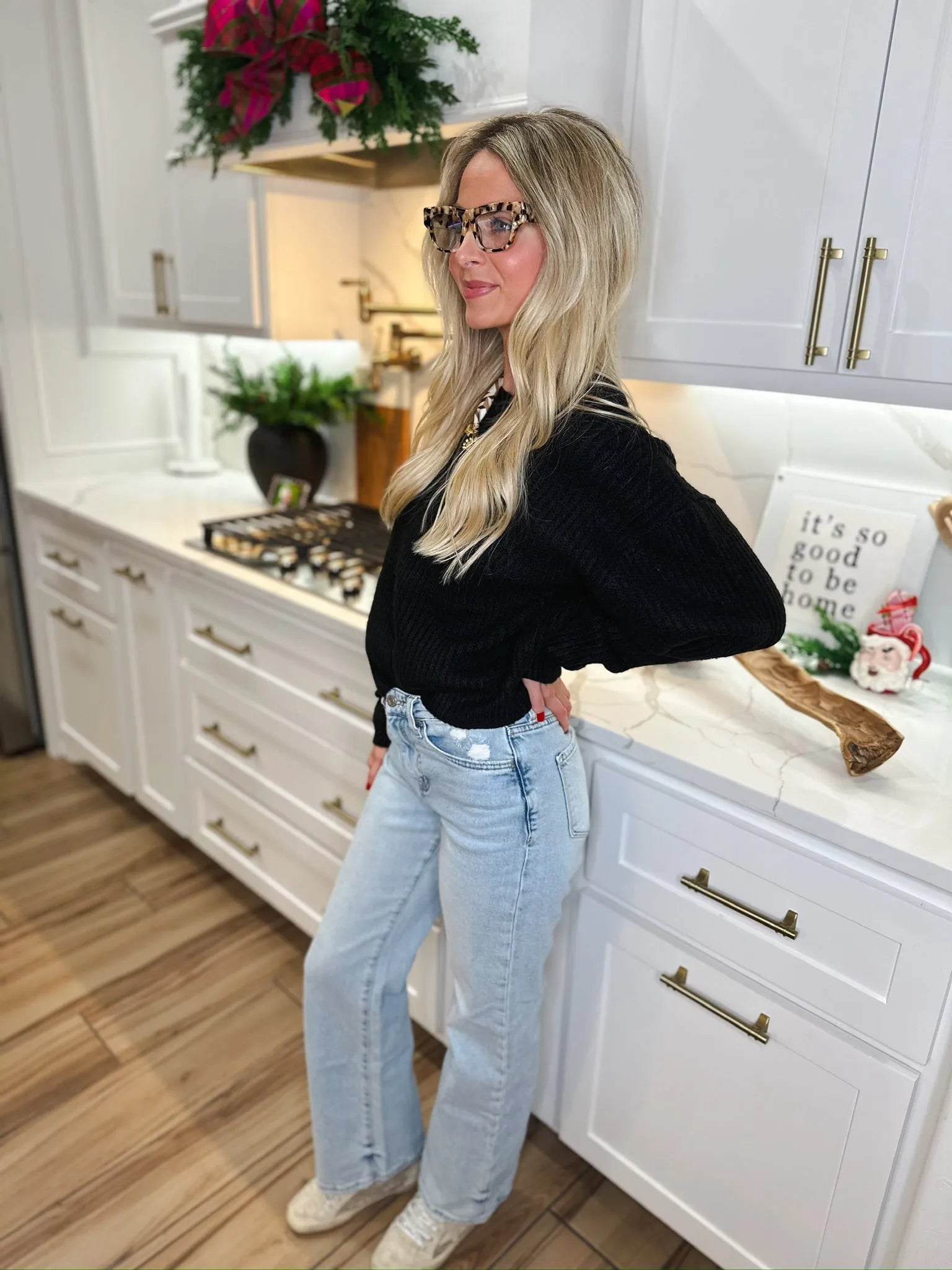 A woman wearing a black ribbed sweater and light wash distressed jeans leans against a kitchen counter.