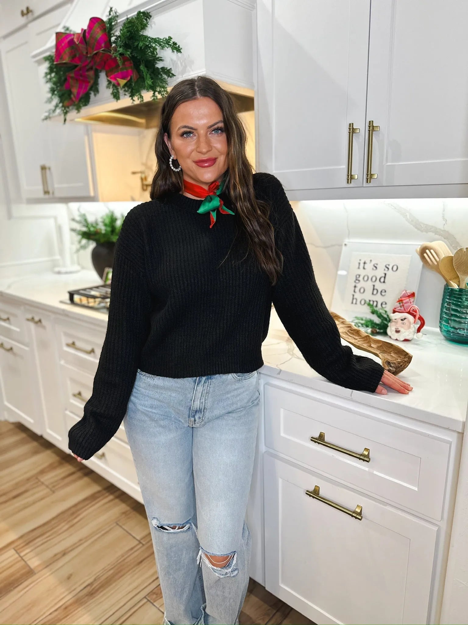 A woman in a black ribbed sweater and light blue ripped jeans poses in a kitchen, wearing a red and green festive scarf.