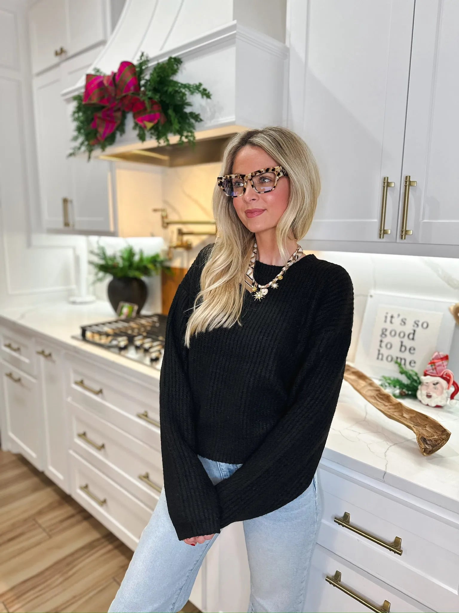 A blonde woman wearing a black ribbed sweater, light wash jeans, statement glasses, and a star pendant necklace stands in a