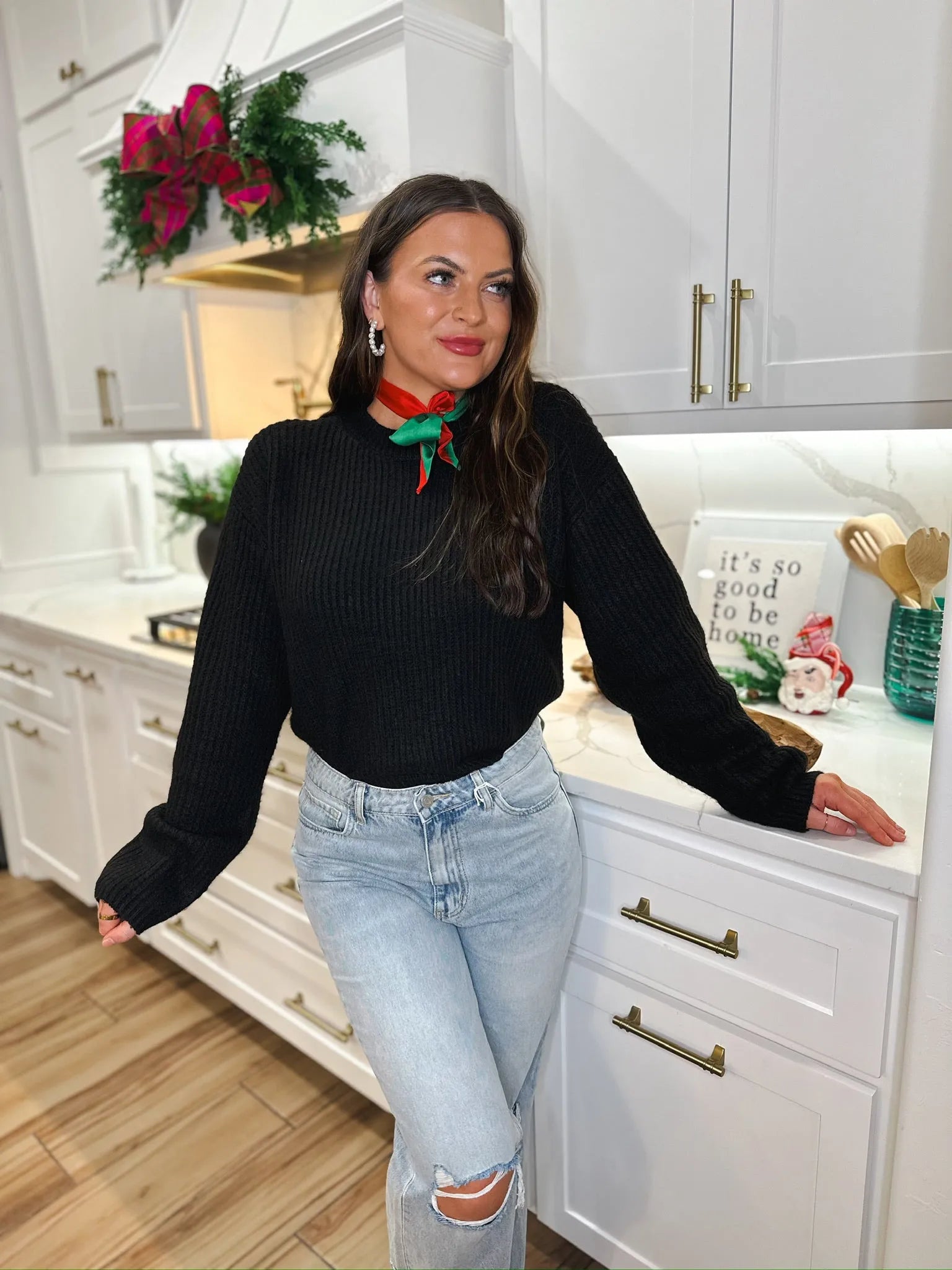 Woman in black ribbed sweater, light wash ripped jeans, and red and green holiday scarf in a modern kitchen.