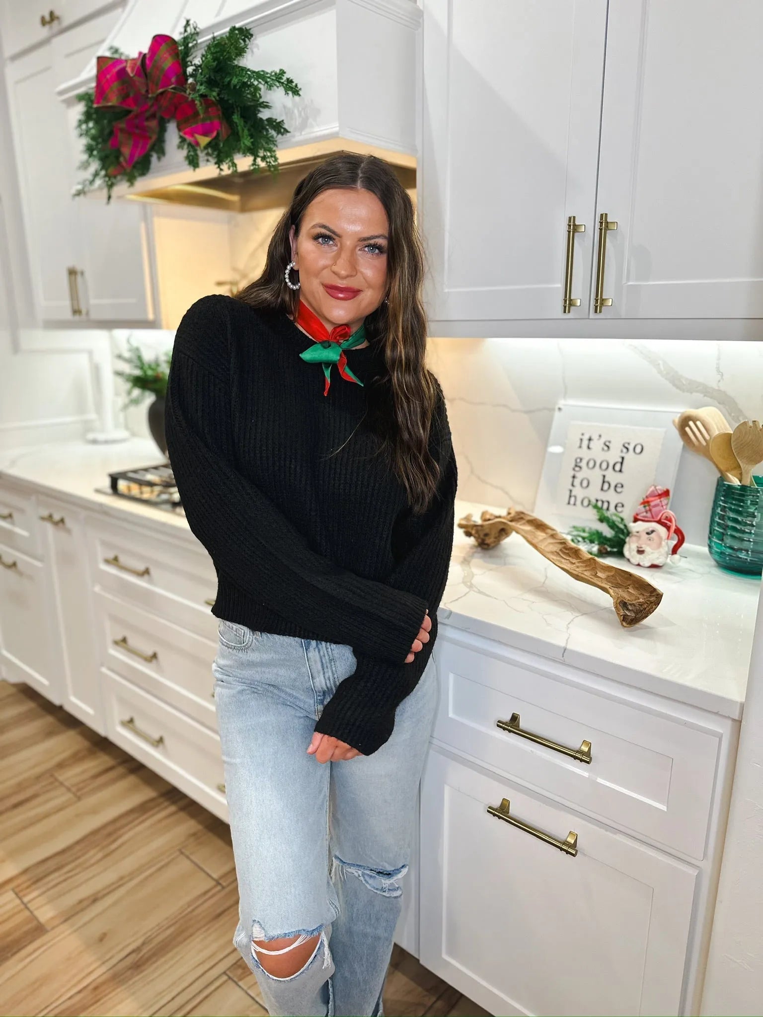 Woman wearing a black ribbed sweater, light wash distressed jeans, and a festive red and green scarf tied around her neck