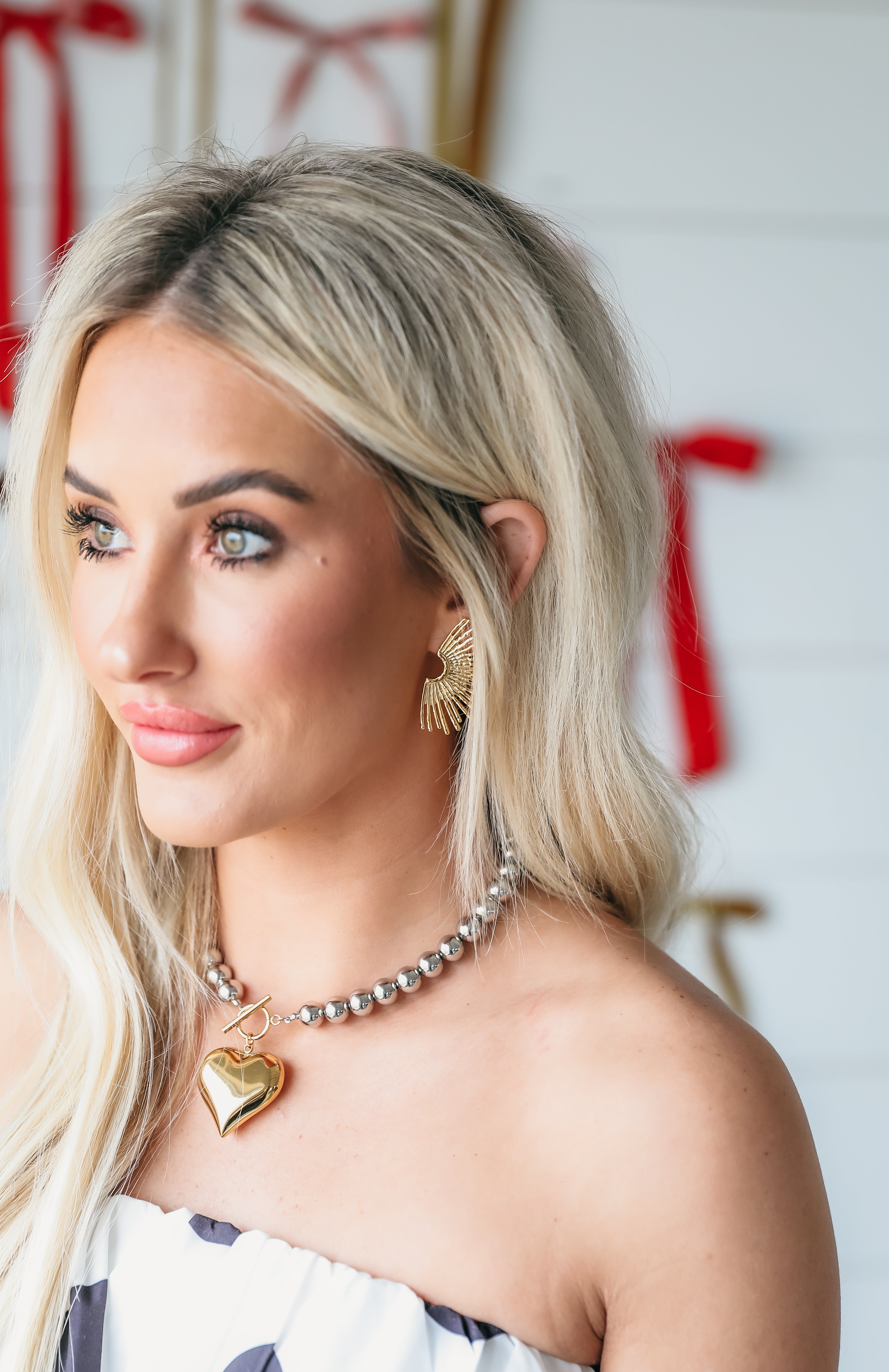 A woman wearing a two-tone heart necklace and gold fan earrings. The necklace features a large gold heart pendant on a
