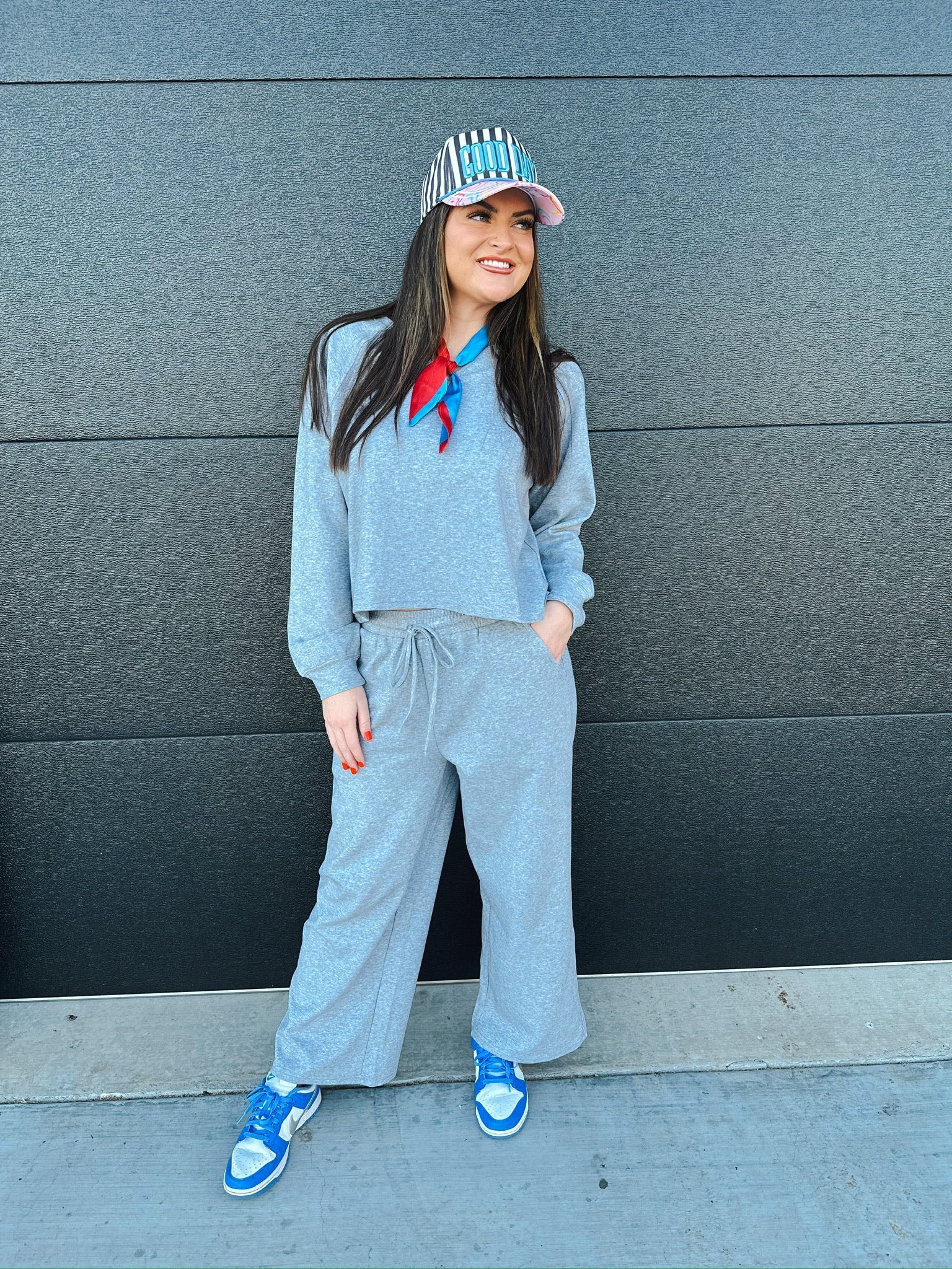Woman wearing a heather grey V-neck cropped sweatshirt and wide-leg sweatpants with a red and blue scarf and striped cap.