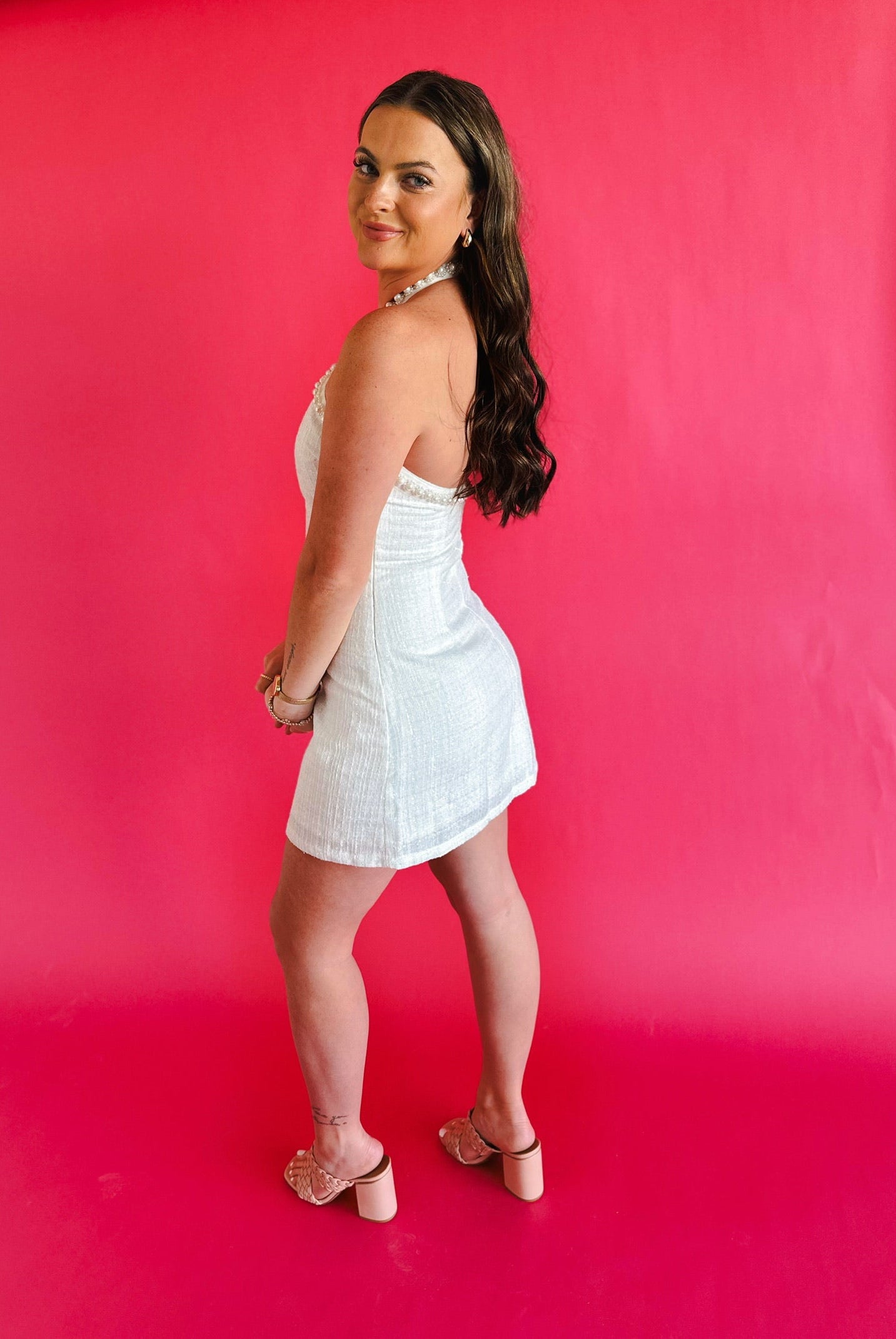 Woman in a white tweed halter mini dress with pearl trim, standing against a bright pink background. She wears gold hoop