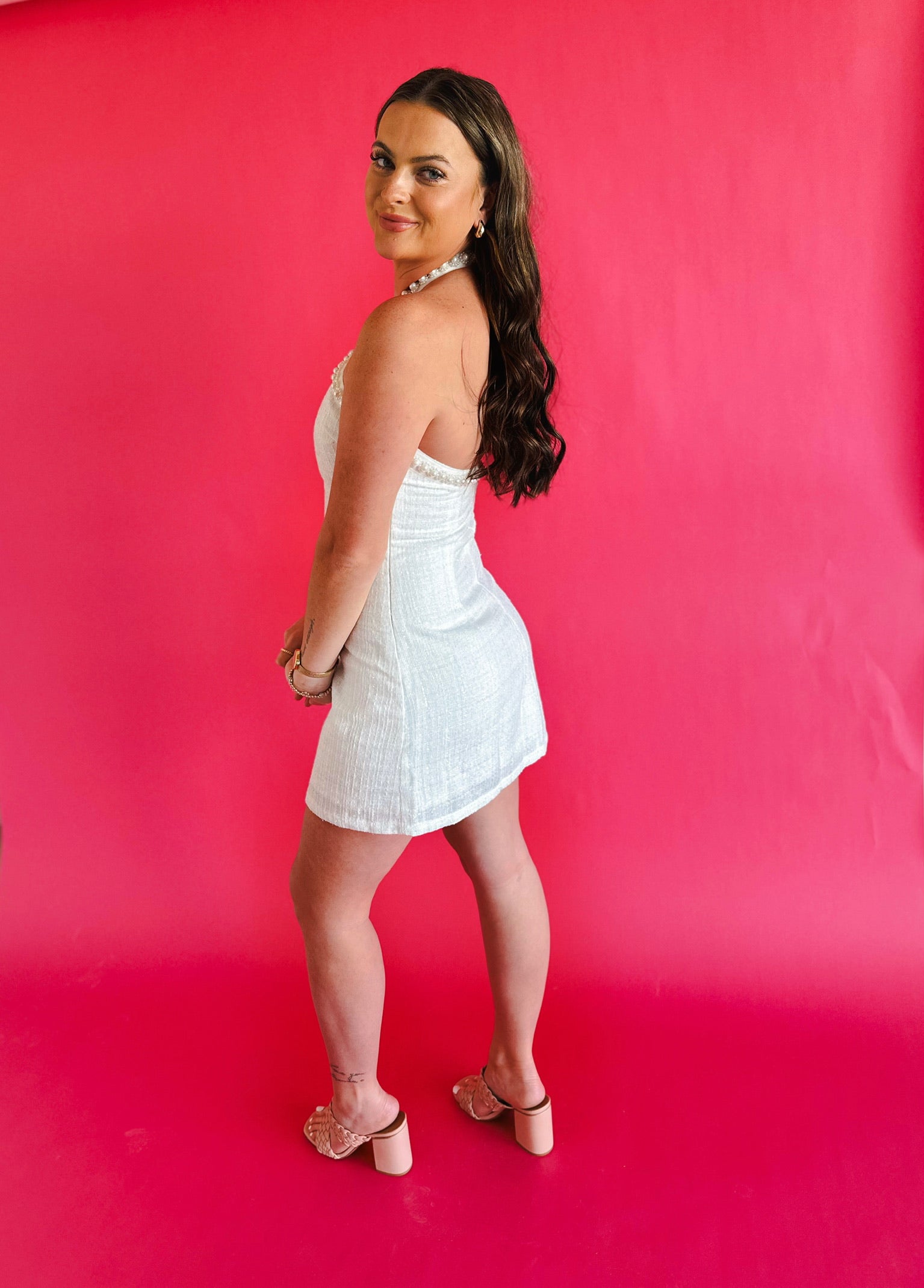 Woman in a white tweed halter mini dress with pearl trim, standing against a bright pink background. She wears gold hoop