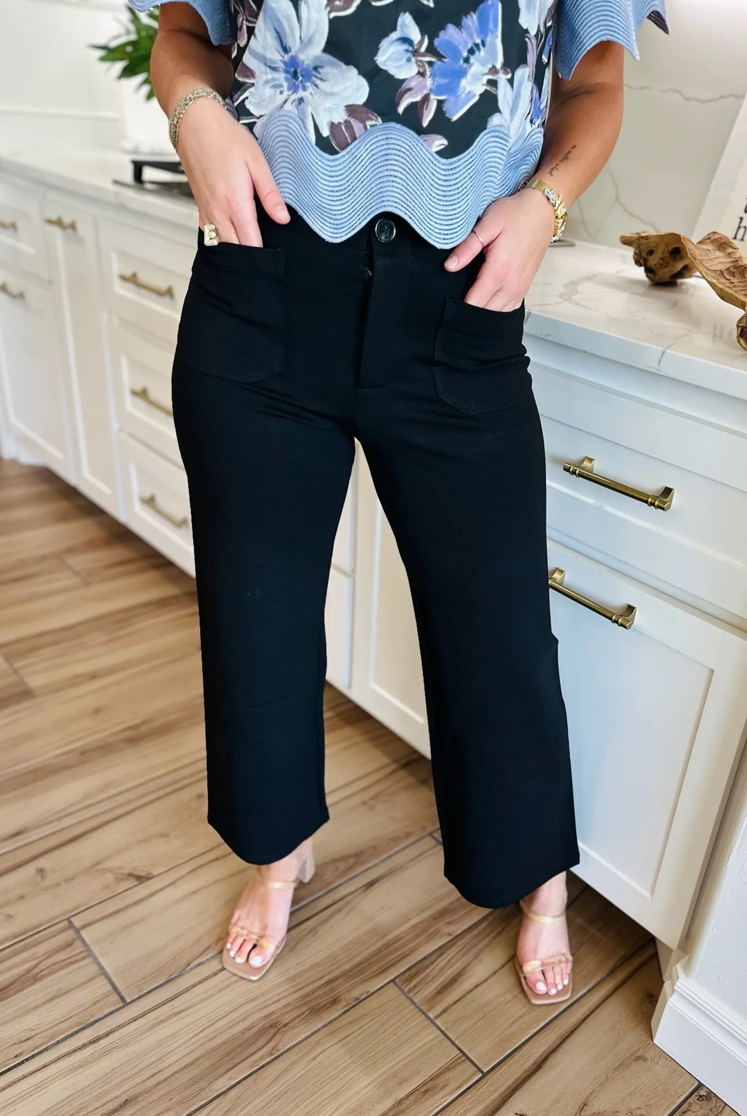 Black ponte wide leg pants with front pockets and button closure, worn by a woman with a floral top and gold sandals.