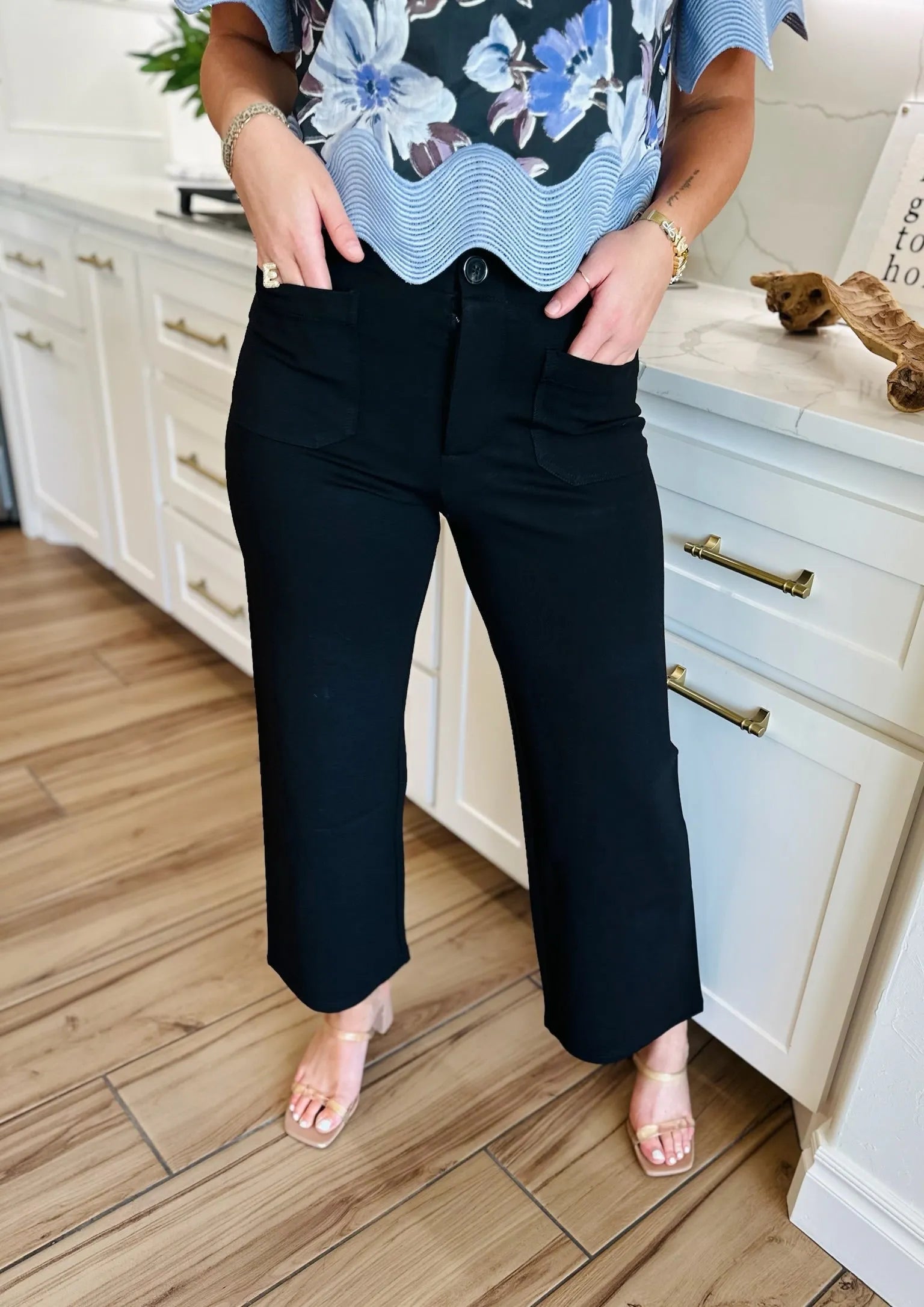 Black ponte wide leg pants with front pockets and button closure, worn by a woman with a floral top and gold sandals.