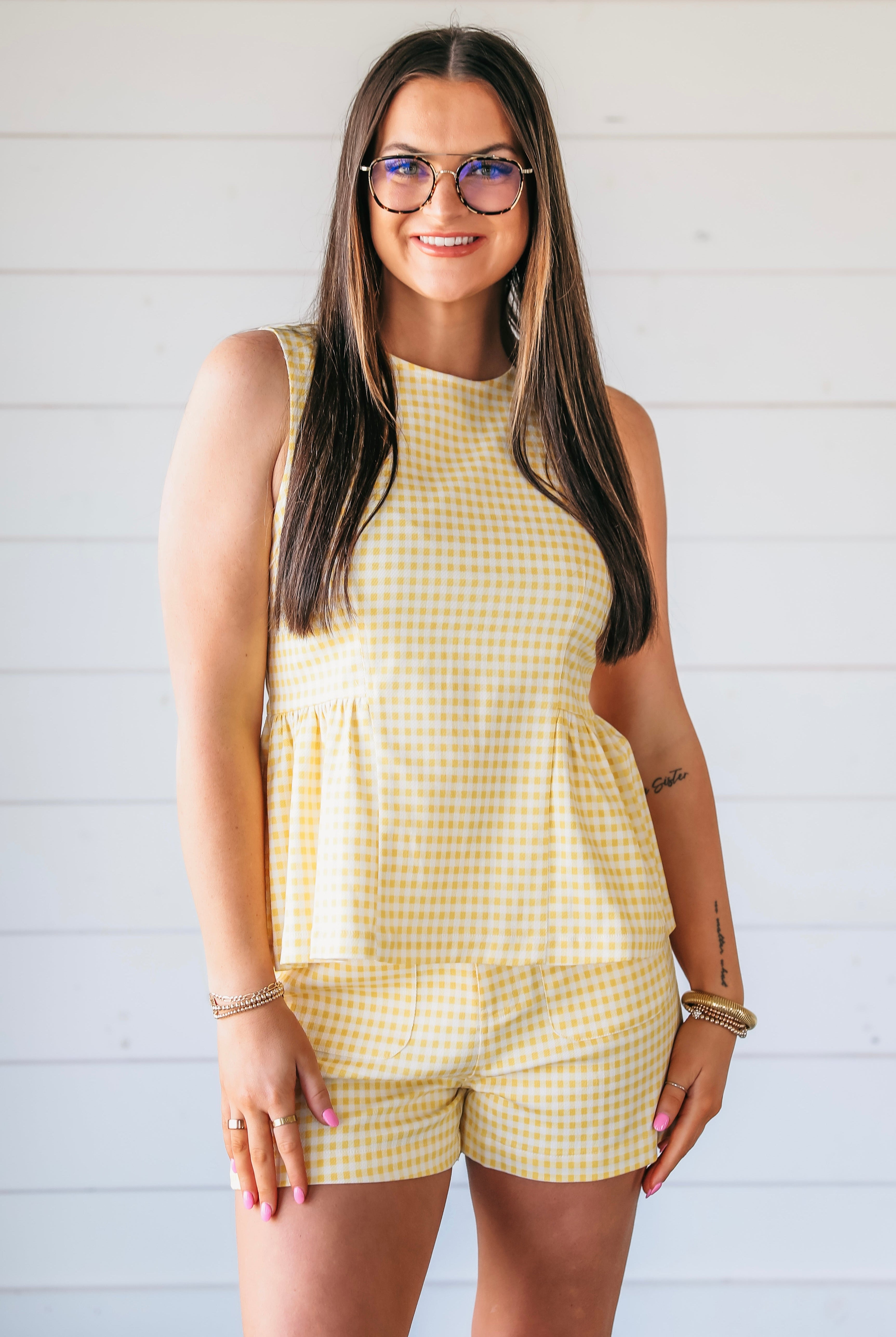Woman wearing a yellow gingham peplum top and shorts set. She has on glasses and bracelets.