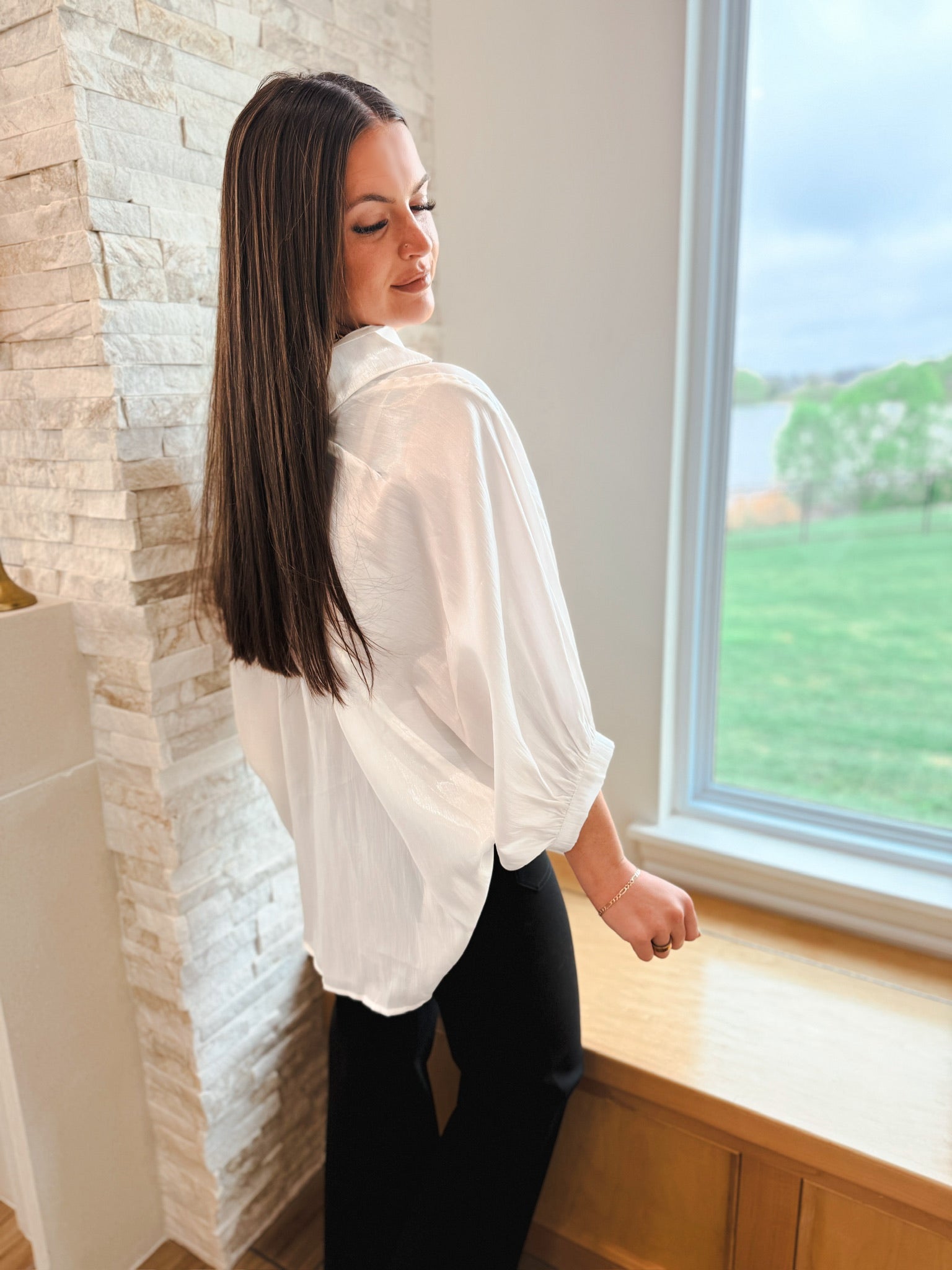 Woman wearing a white shiny button-front blouse with wide, long sleeves and black pants, standing by a window.