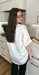 Woman from behind wearing an ivory satin v-neck high-low blouse with cuffed short sleeves. She has long brown hair and