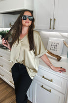 Woman wearing a taupe satin V-neck blouse and black pants, leaning on a kitchen counter.