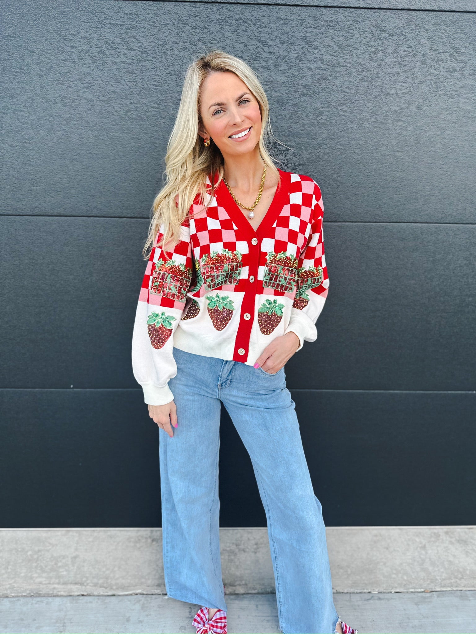 Woman wearing Queen of Sparkles red and white checkered sequin strawberry cardigan with light wash wide-leg jeans.