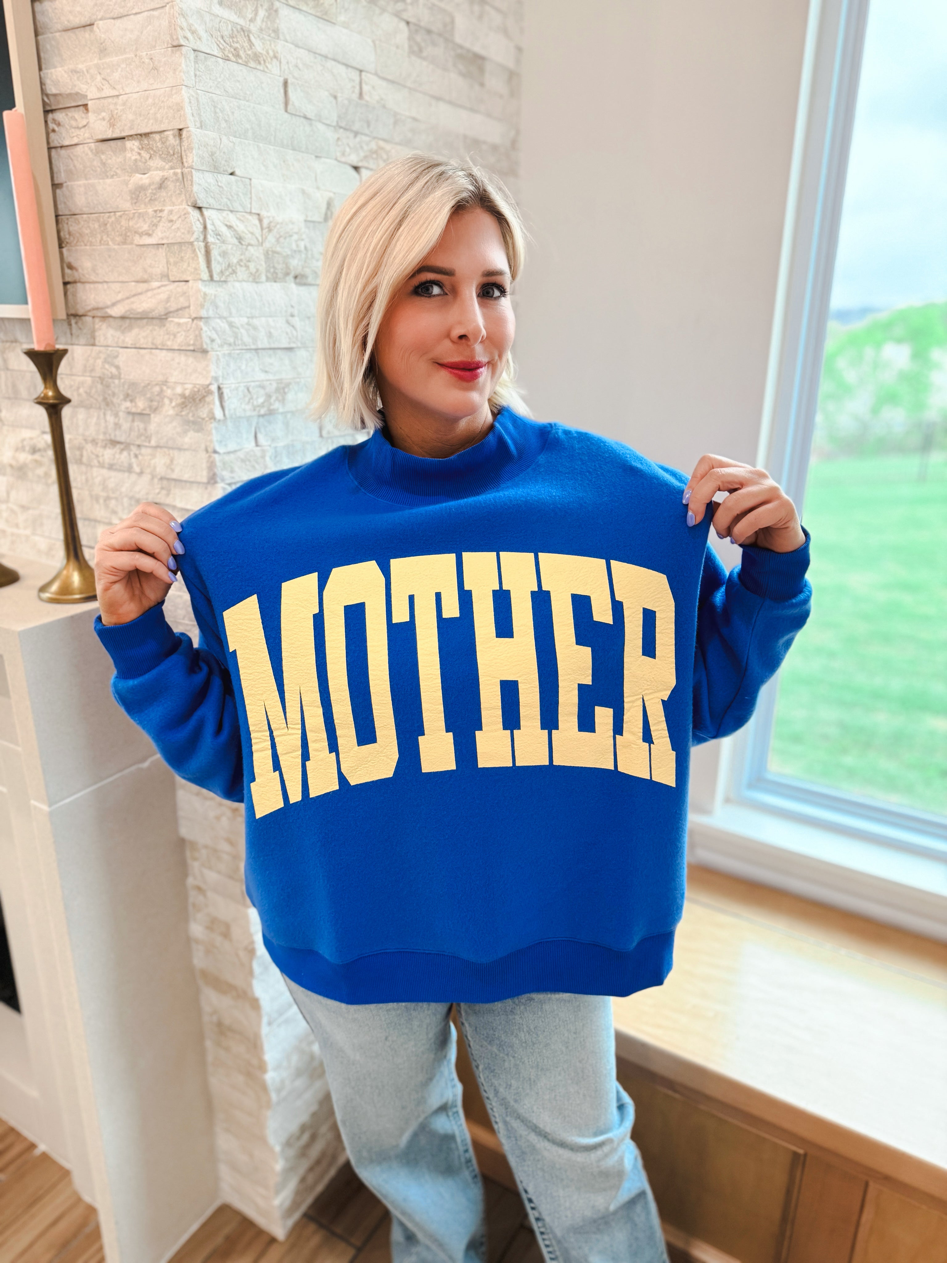 Royal blue mockneck oversized sweatshirt with yellow "MOTHER" graphic print on front. Worn by a blonde woman.