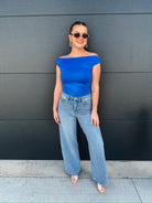Woman wearing light wash, slouch wide-leg jeans and a bright blue off-the-shoulder top, sunglasses, and hoop earrings.
