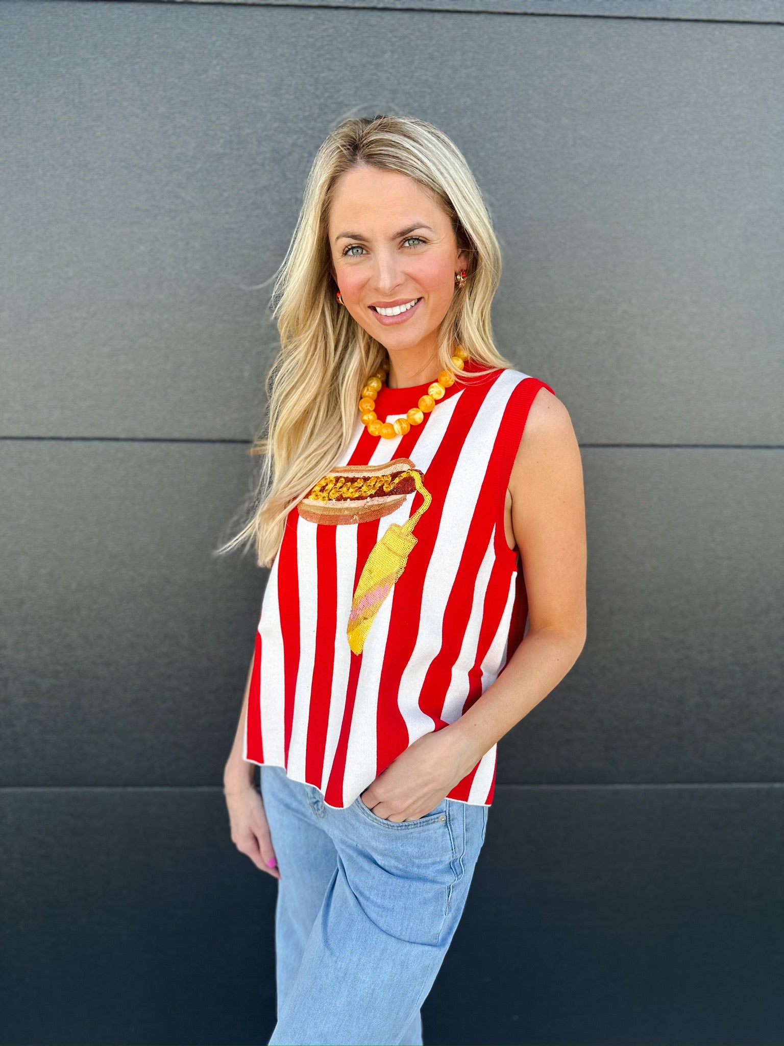 Woman wearing a red and white striped knit tank with a sequined hotdog graphic. Light wash jeans and yellow beaded necklace.