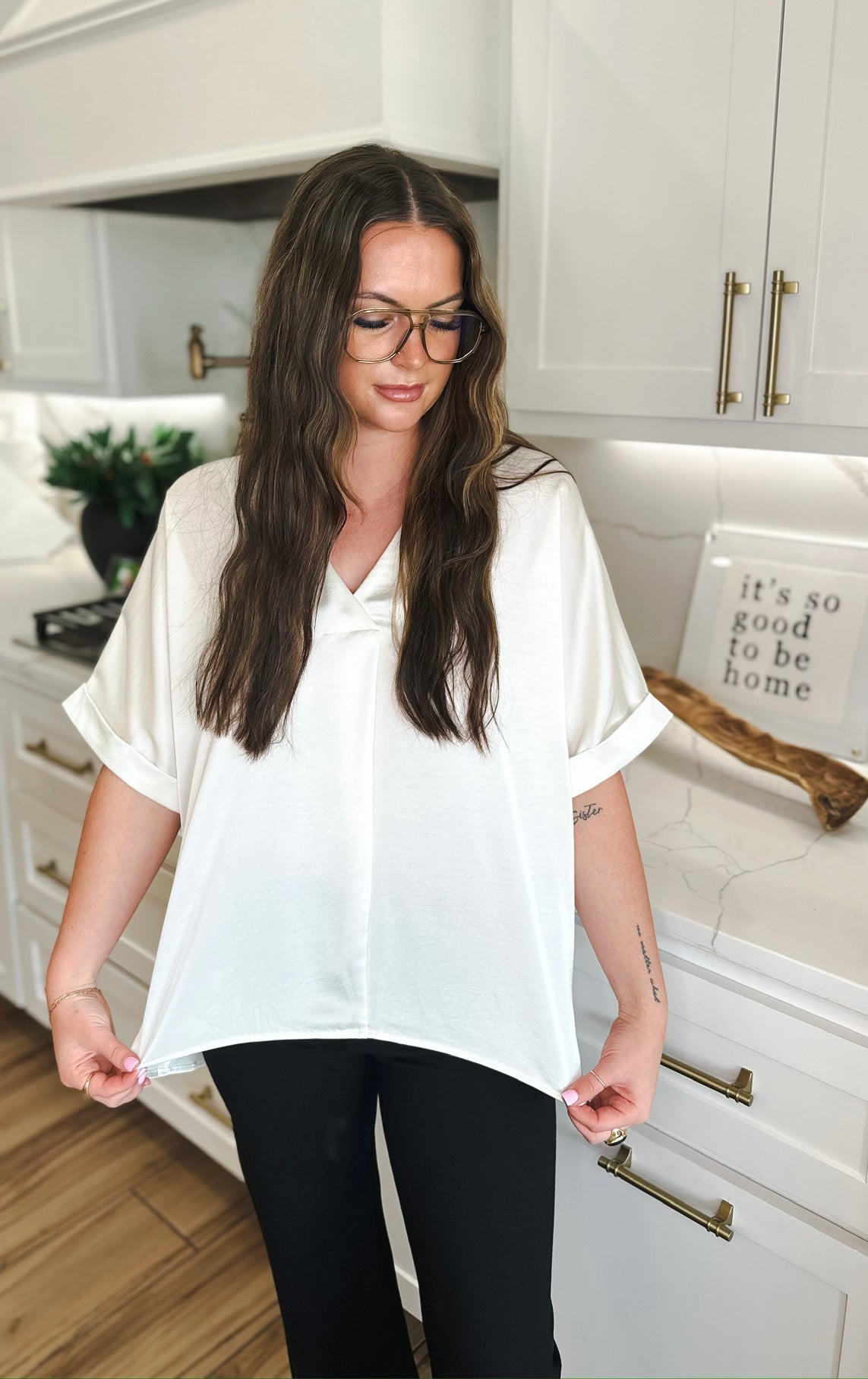 Woman wearing an ivory satin V-neck high-low blouse and black pants, styled casually in a kitchen.