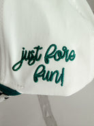 White caddie hat with "just fore fun!" embroidered in green script. Herringbone pattern visible on brim.