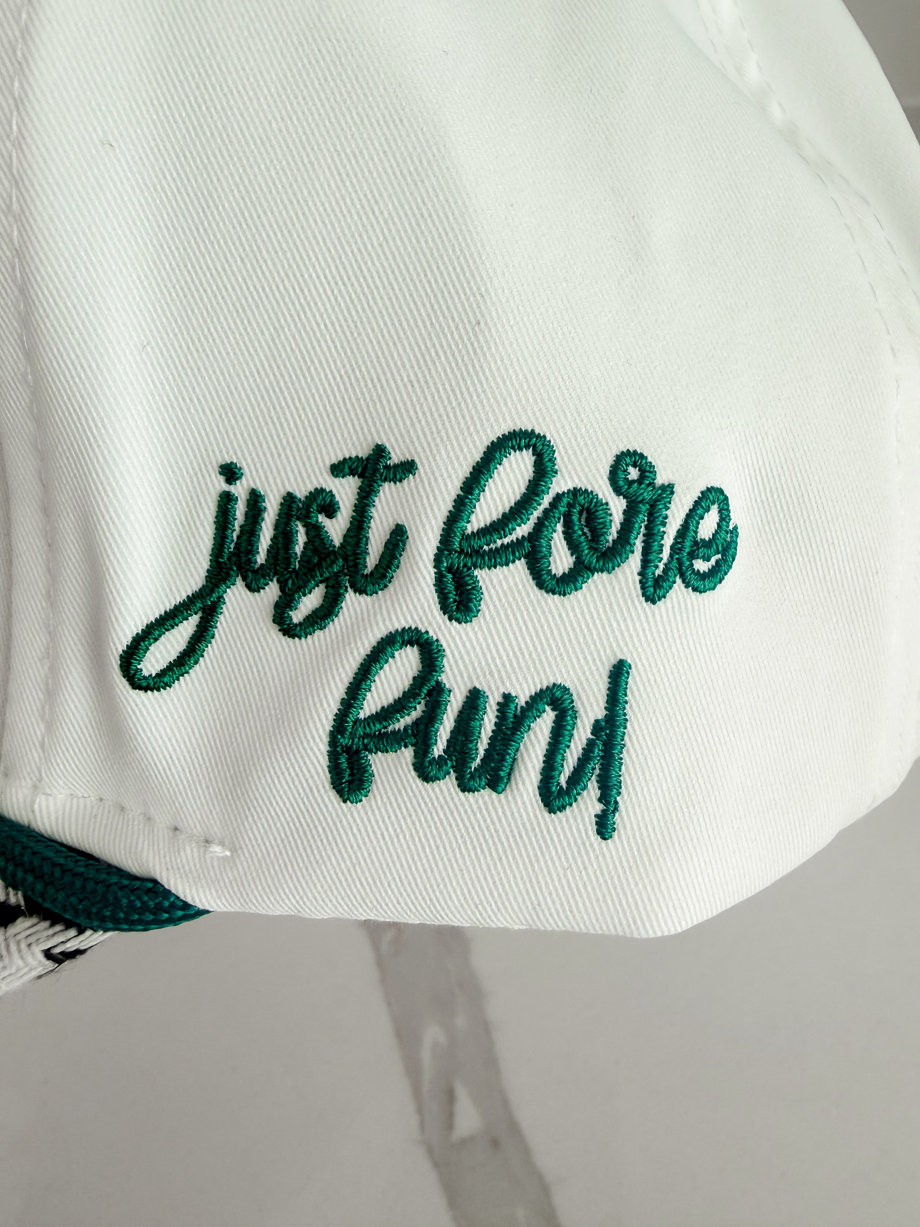 White caddie hat with "just fore fun!" embroidered in green script. Herringbone pattern visible on brim.