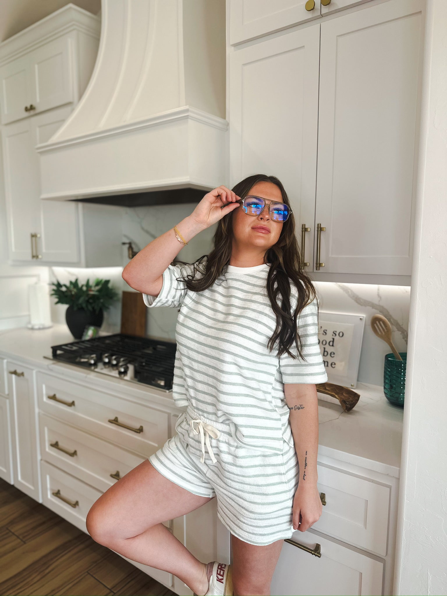 Woman wearing a white and sage green striped lounge set with shorts and a t-shirt, holding her glasses.