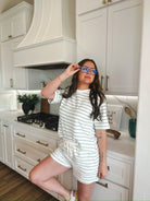 Woman wearing a white and sage green striped lounge set with shorts and a t-shirt, holding her glasses.