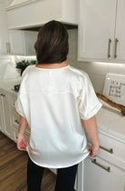 Back view of a woman wearing an ivory satin V-neck high-low blouse with cuffed short sleeves.