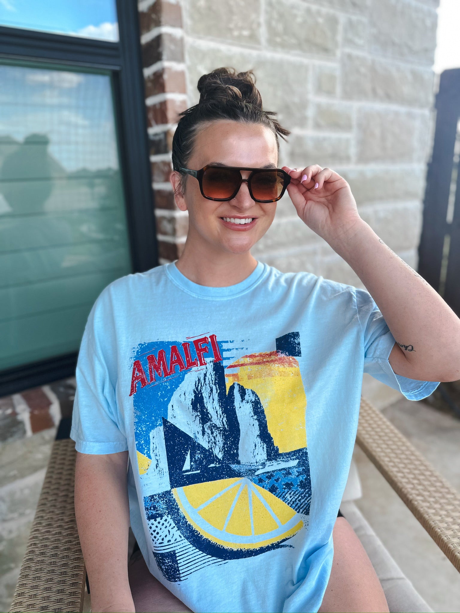 Woman in a light blue oversized tee with a graphic print of Amalfi Coast cliffs, a lemon slice, and a sailboat. She wears