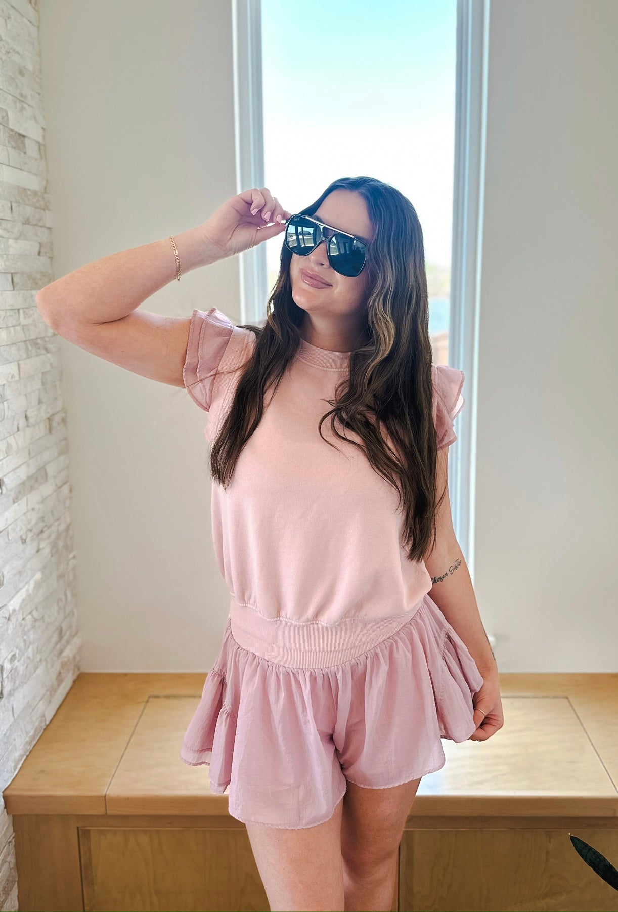 Woman wearing a light pink athleisure set with ruffle sleeve pullover and crinkle nylon shorts. She is wearing sunglasses.