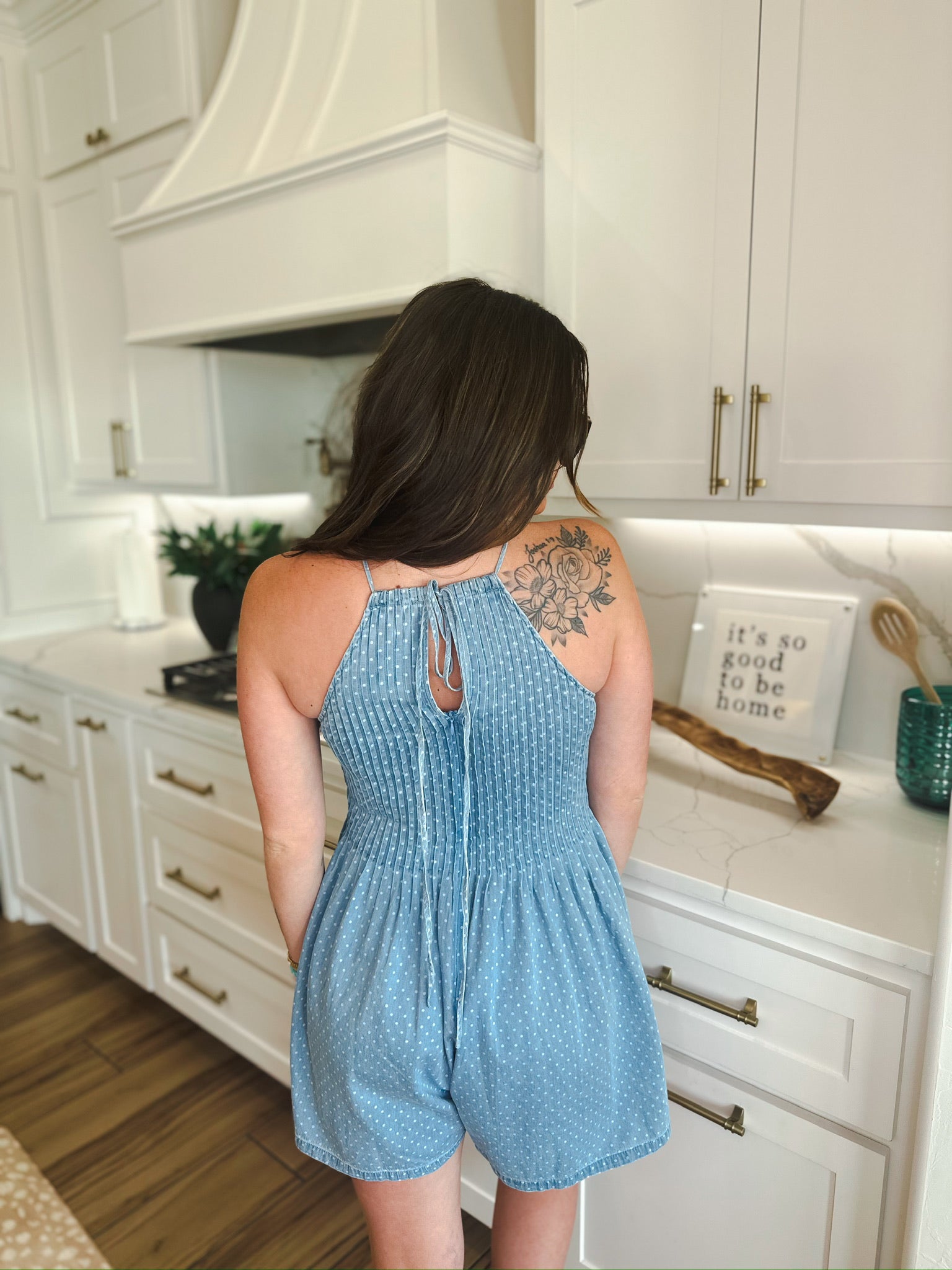 Back view of a woman wearing a light blue denim romper with white polka dots, featuring a halter neck and a tie-back detail.