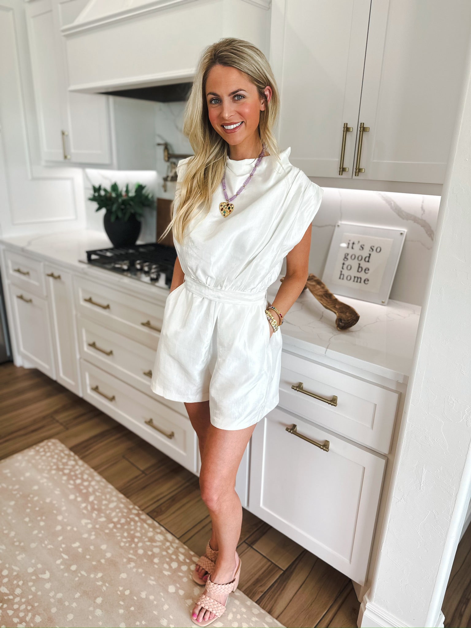 Woman in white shimmer romper with cowl neck and cap sleeves. Romper has a belted waist and pleated shorts.