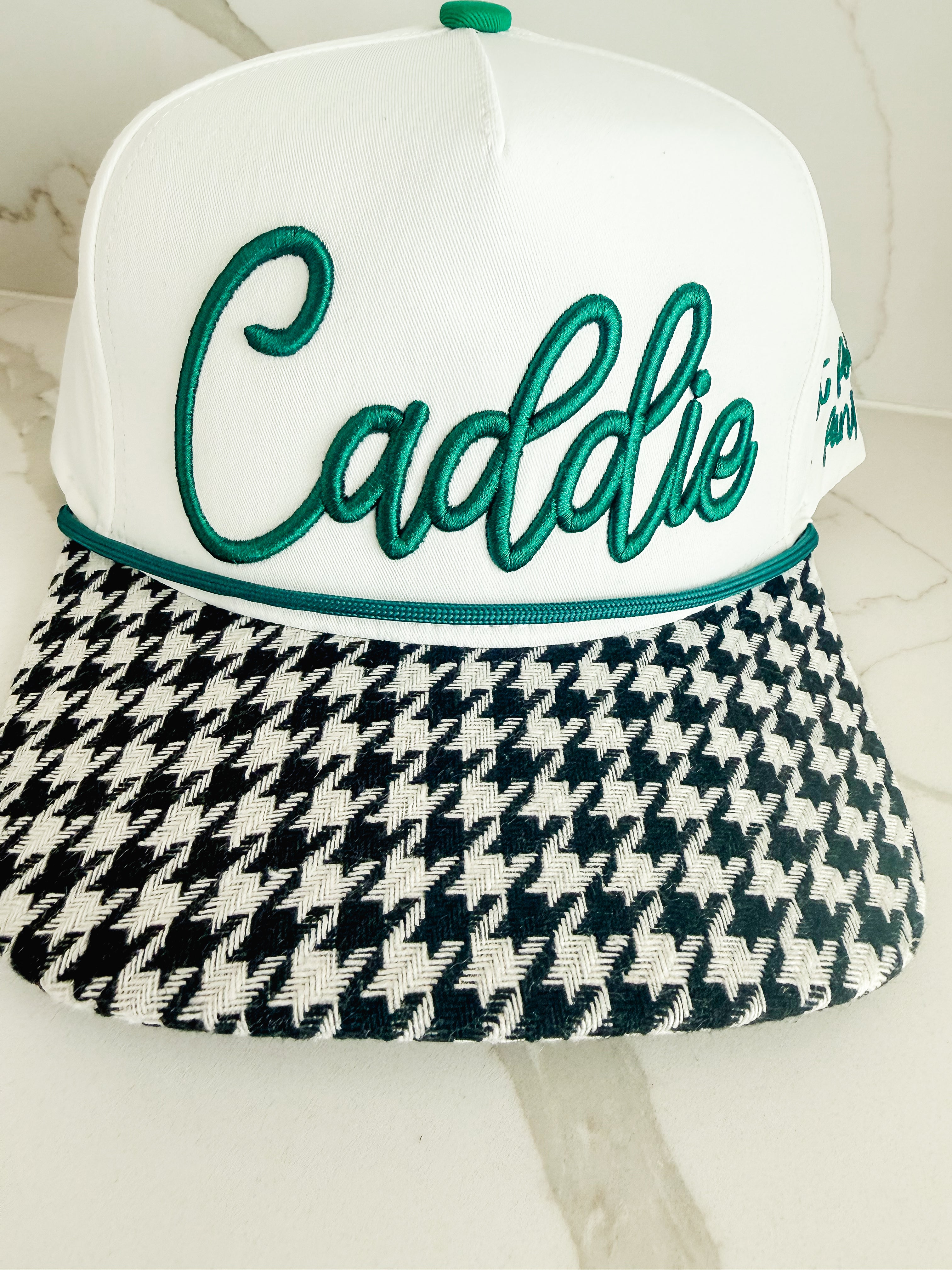 White caddie hat with green embroidered script logo and black and white houndstooth brim. Green rope accent above brim.