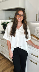 Woman wearing an ivory satin V-neck high-low blouse and black pants, smiling and standing in a kitchen.