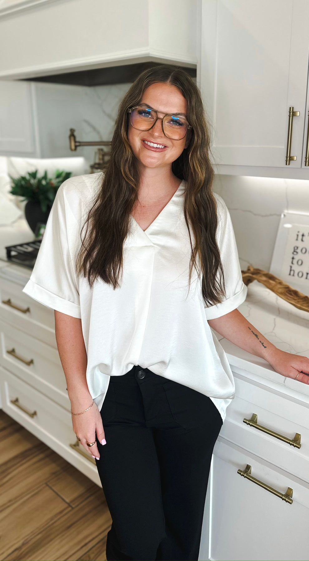 Woman wearing an ivory satin V-neck high-low blouse and black pants, smiling and standing in a kitchen.