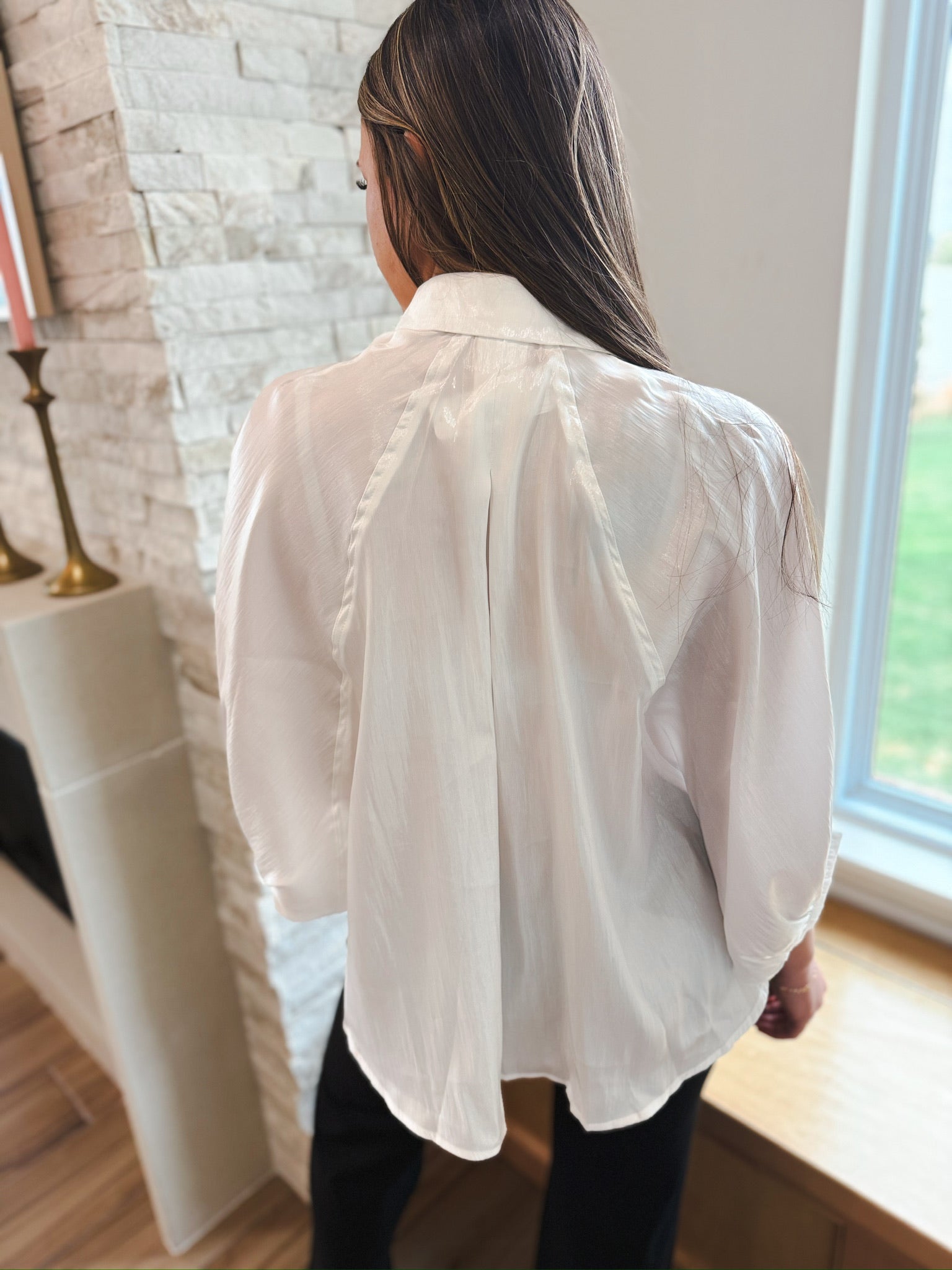 Rear view of a white shiny button-front blouse with wide, long sleeves and a textured fabric. The top has a central back