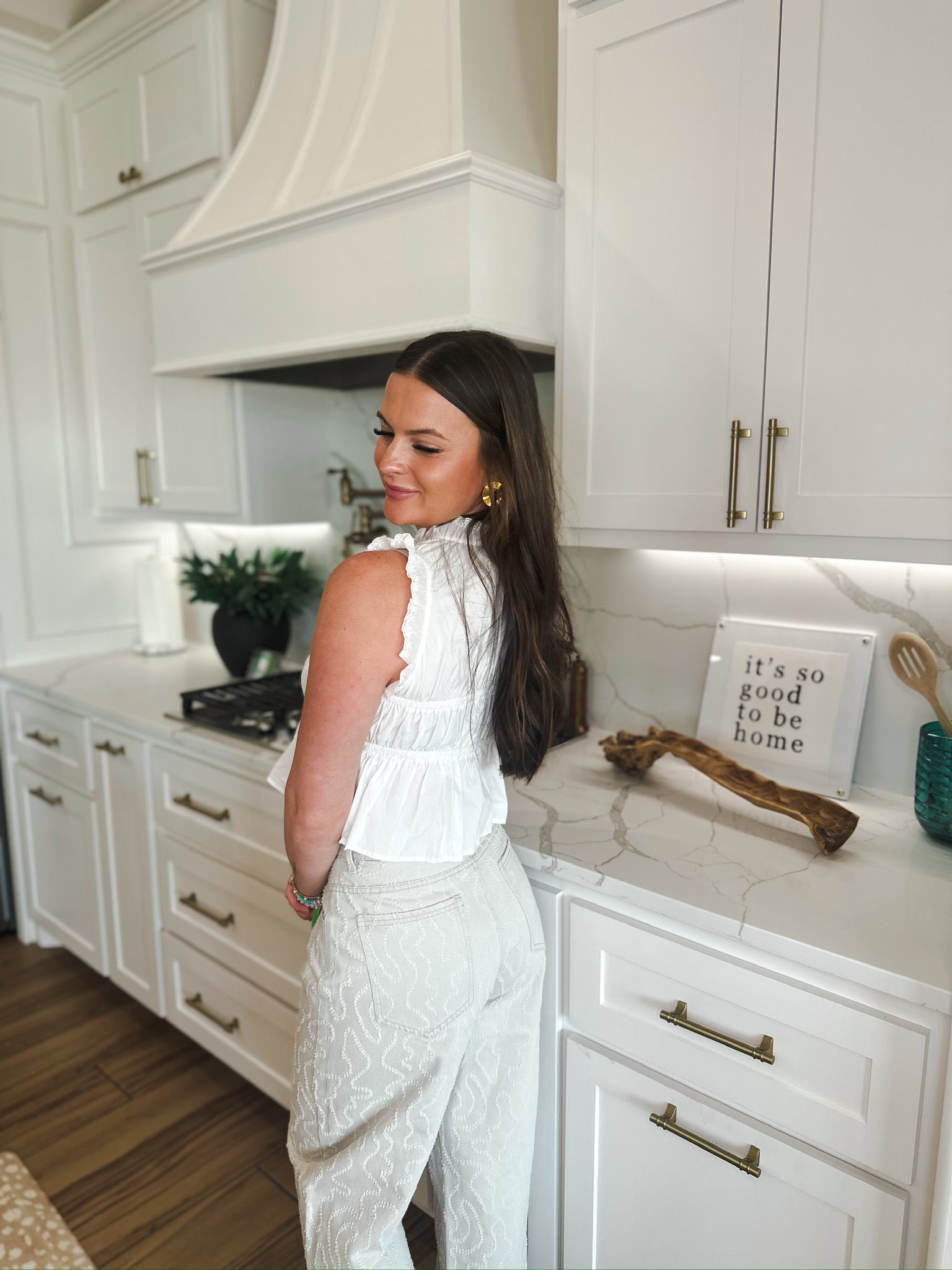 Woman in a white lace trim tie-front peplum top and light-wash patterned pants, standing in a modern white kitchen.