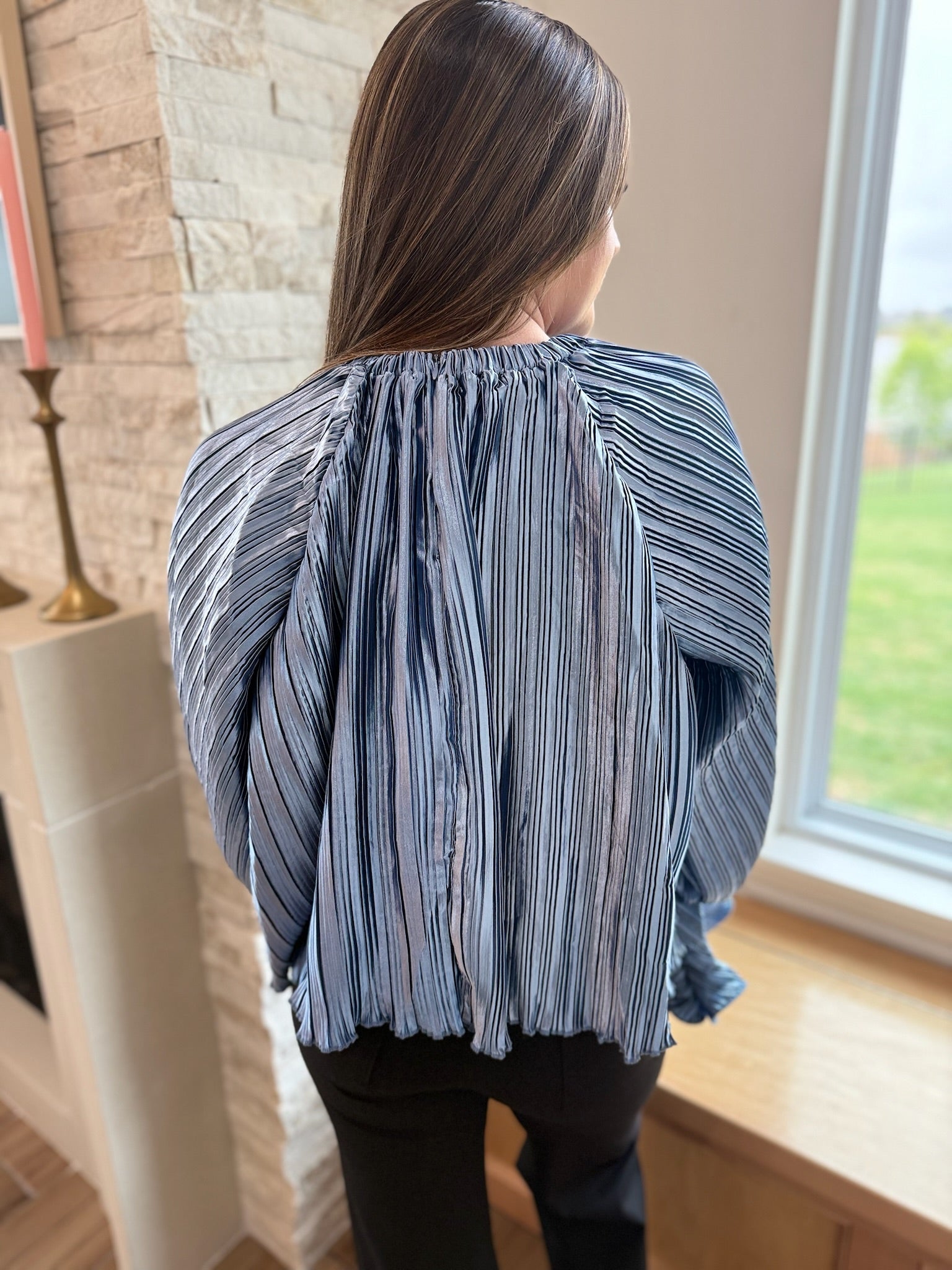 Back view of a woman wearing a slate blue pleated long sleeve top with vertical stripes and ruffled cuffs and hem.