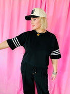 Woman wearing a black short-sleeve top with white stripes on the sleeves, black drawstring pants, and a beige baseball cap