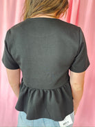 Back view of a black peplum jacket with short sleeves, featuring a gathered hemline and jeweled button detail on the upper