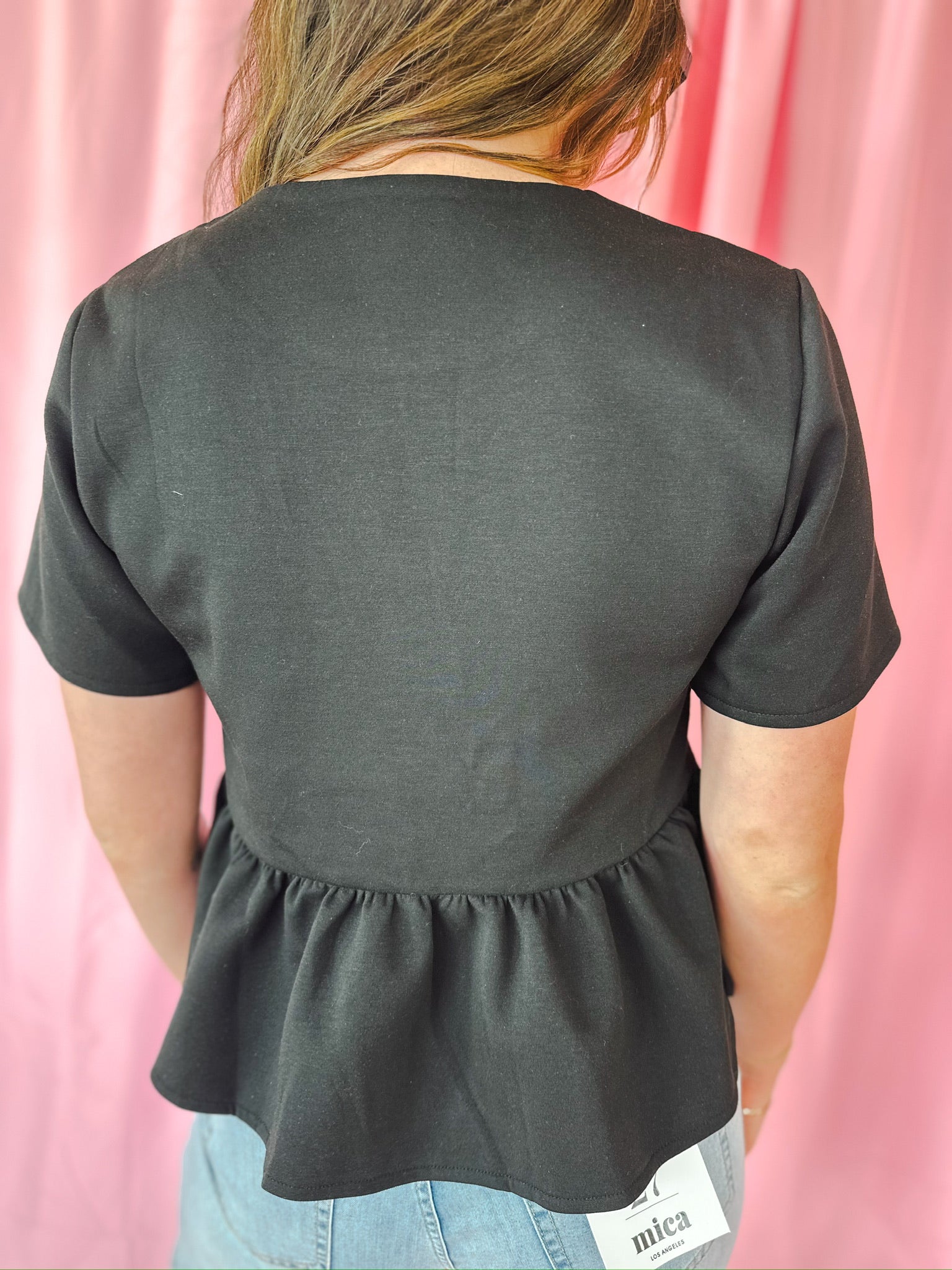 Back view of a black peplum jacket with short sleeves, featuring a gathered hemline and jeweled button detail on the upper